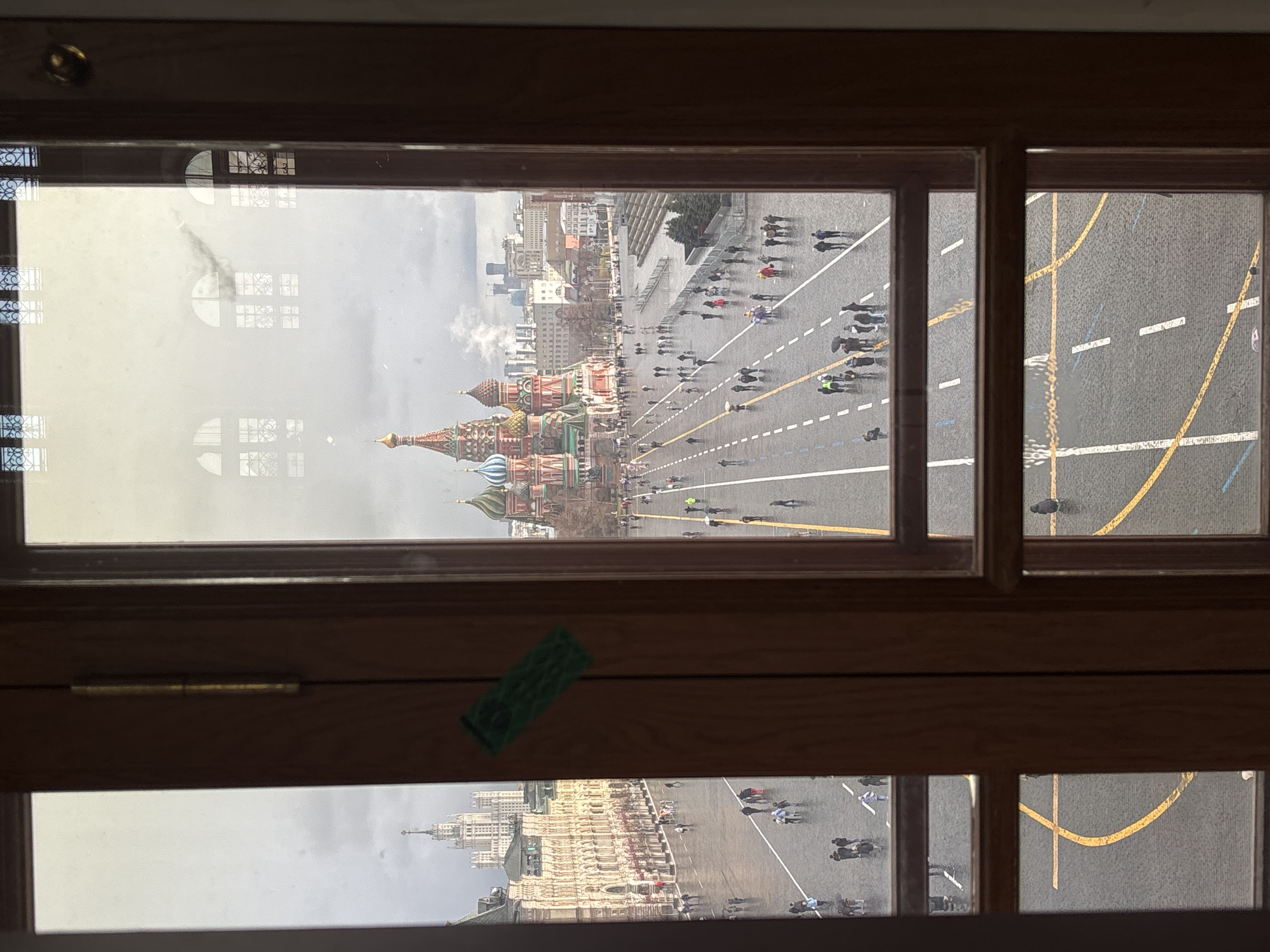 The image is of a view of the Red Square in Moscow, Russia as seen through a window. The window is composed of wooden frames and panes. Through the top left pane, one can see the iconic domes of Saint Basil's Cathedral and a wide, gray plaza filled with people. The top right pane shows a portion of the square with yellow lines painted on the asphalt. A green sticker is attached to the wooden frame separating the top and bottom panes of the window. The bottom panes of the window present an inverted but similar view of the Red Square, with buildings and people visible. The environment appears to be overcast.