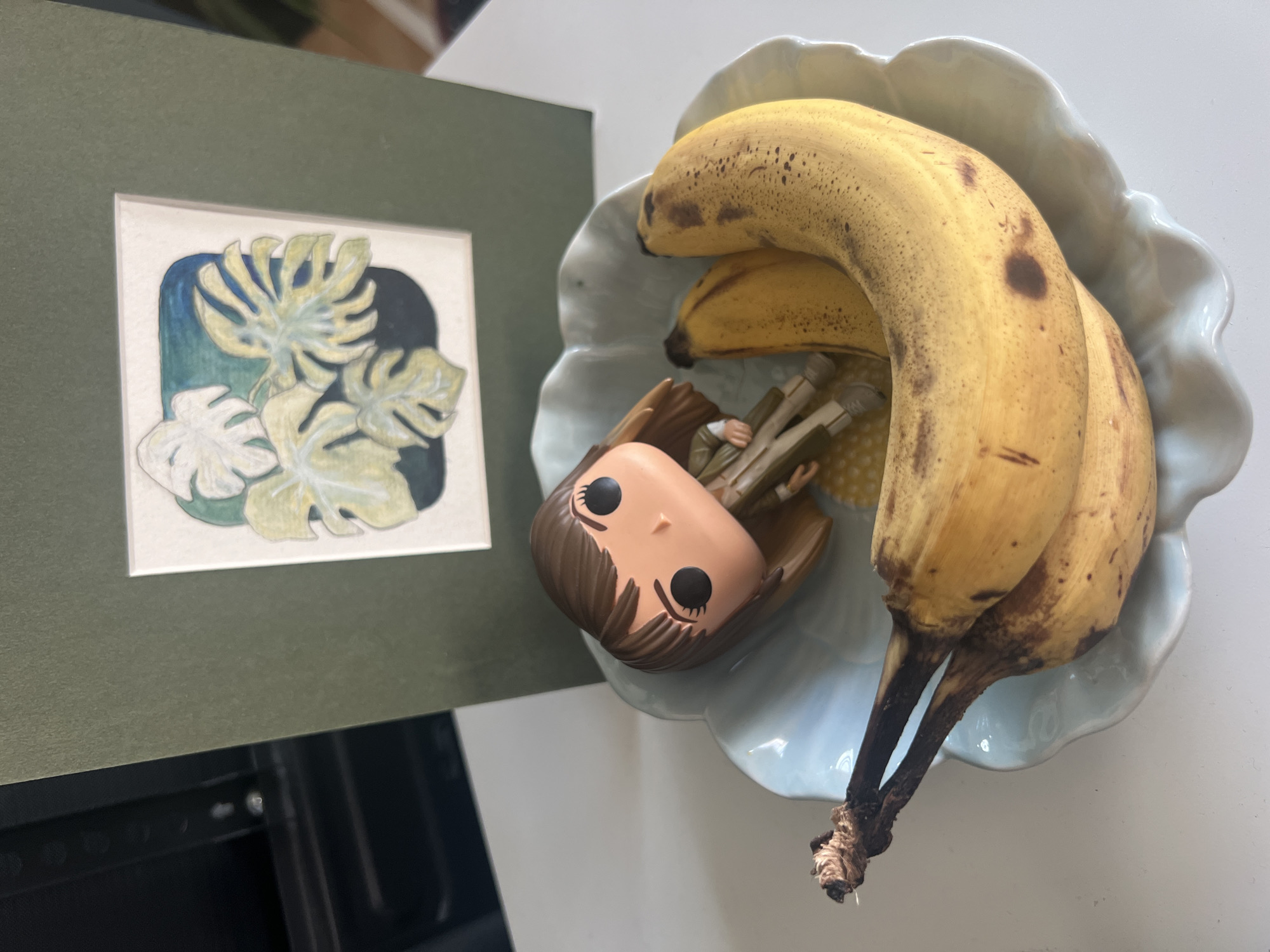 The image depicts a shallow, light blue, scalloped-edge bowl containing two bananas and a figurine. The bananas are ripe, with yellow skin and brown spots. The figurine, which appears to be a Funko Pop character, is positioned on top of the bananas, with its head tilted back. Behind the bowl is a dark green mat or frame, within which is a small watercolor painting of monstera leaves. The overall scene is set on a white surface, likely a table. The image does not contain any information that can be used to determine its location in the city of Mollet del Vallès, Spain.