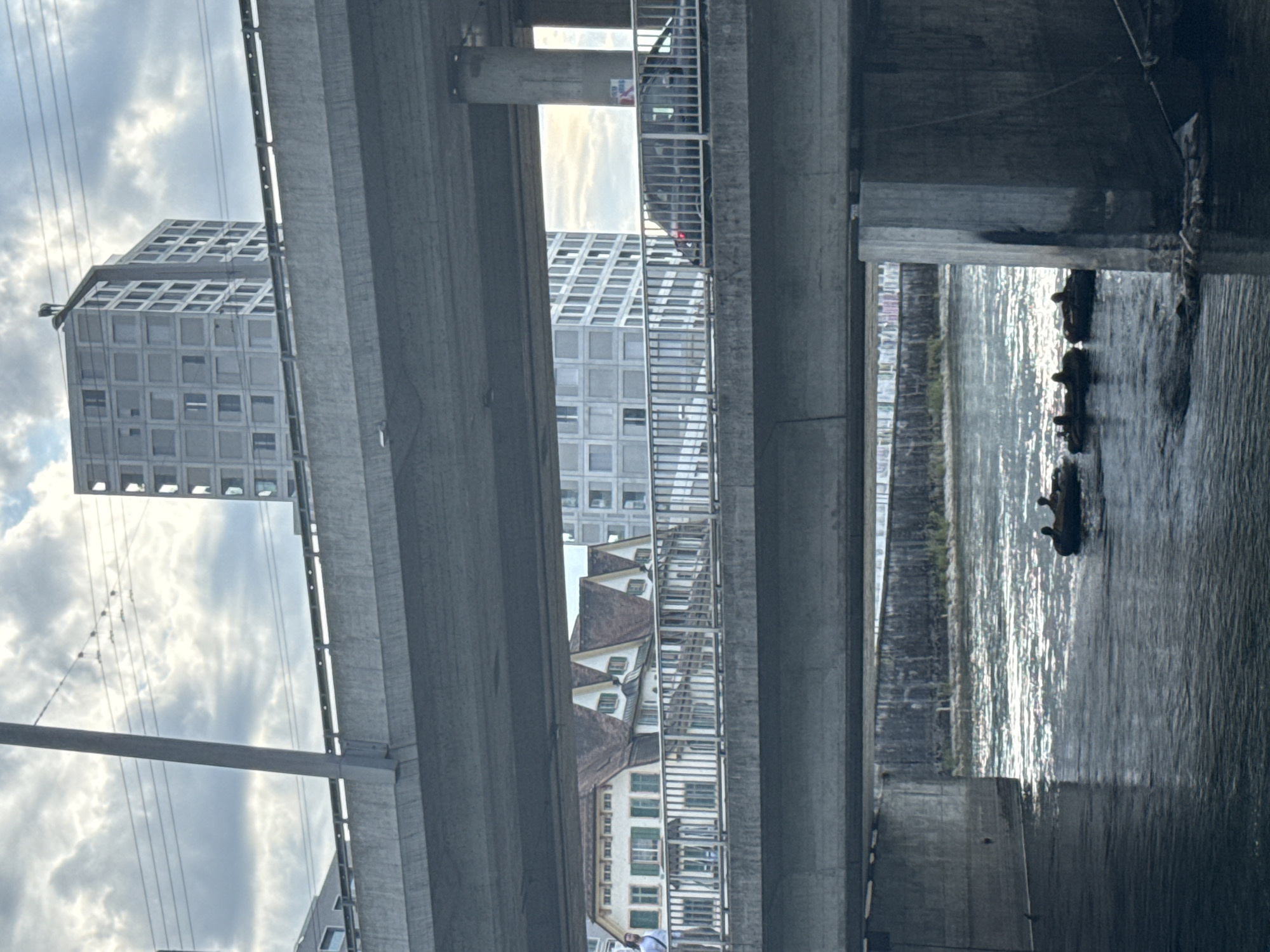This image captures a view under a multi-level bridge system in Zurich, Switzerland. In the foreground, a river flows, reflecting the sunlight. Several inflatable tubes, each carrying one or two individuals, are navigating the river downstream.
Above the river, a lower bridge deck is visible, supported by concrete pillars. Beyond this, a street level bridge is seen, with a railing along its edge. A man is walking on this bridge, and a dark-colored car is driving across it. In the background, a row of houses with distinctive peaked roofs is visible, followed by modern, multi-story buildings. The sky is overcast with scattered clouds. The overall impression is an urban landscape with elements of both historical and contemporary architecture, integrated with natural elements like the river.
