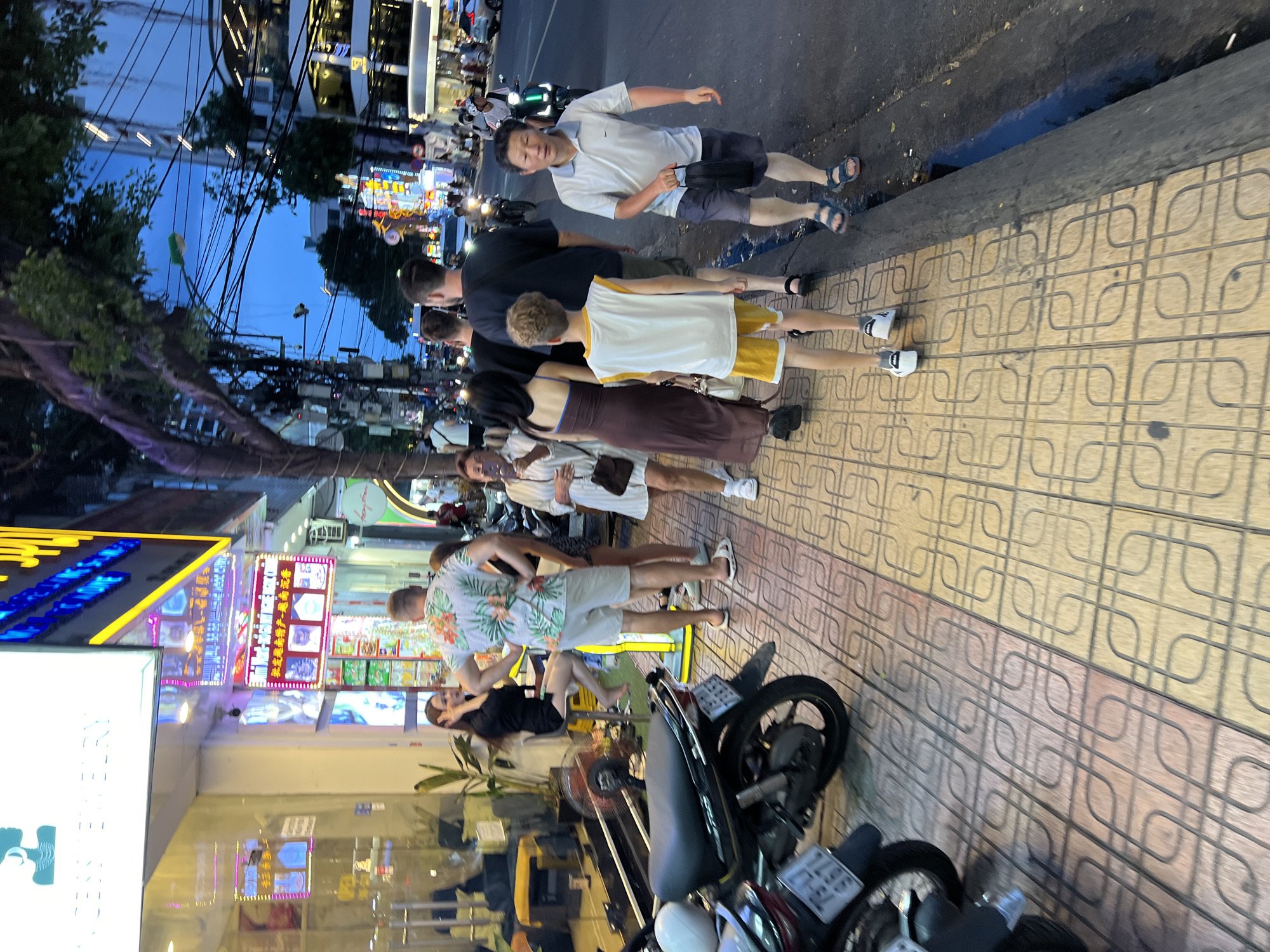 The image captures a street scene in Nha Trang, Vietnam, likely during the evening. The foreground features a sidewalk with patterned yellow and red tiles, bordered by a street with a dark, wet edge. Two motorcycles are parked on the left side of the sidewalk.
Several people are walking on the sidewalk. In the center, a young boy in a white tank top and yellow shorts walks ahead of an adult man wearing a light blue polo shirt and dark shorts, holding a black bag. To their left, a woman in a long, dark brown dress walks away from the camera, followed by a woman in a white dress. Further back, a man in a floral print shirt and light shorts stands with his hands on his hips, facing away from the camera, near a woman in a black top and skirt sitting on a stool.
The background reveals a commercial street lined with shops and buildings, many adorned with bright neon signs, indicating commercial activity. A tree with dense foliage extends into the frame from the upper left. The sky above is a deep blue, suggesting twilight or evening hours. The overall atmosphere is that of a busy urban environment.