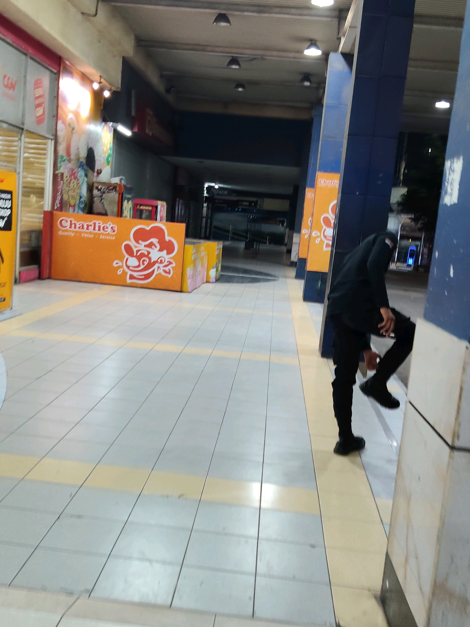 The image captures a person dressed in black, standing with their right leg raised, leaning against a blue pillar. They appear to be adjusting their footwear. The setting is an outdoor walkway with a patterned tiled floor, marked with yellow lines. To the left, there is a vibrant orange display board with the "Charlie's" logo, featuring a cartoon chef, suggesting a food stall or outlet. In the background, a dark corridor is visible, and to the right, a section of a blue and white pillar dominates the foreground. The overall impression is of a commercial area, possibly a pedestrian walkway within a town or city. This specific scene is located in the city of Purwakarta, Indonesia.