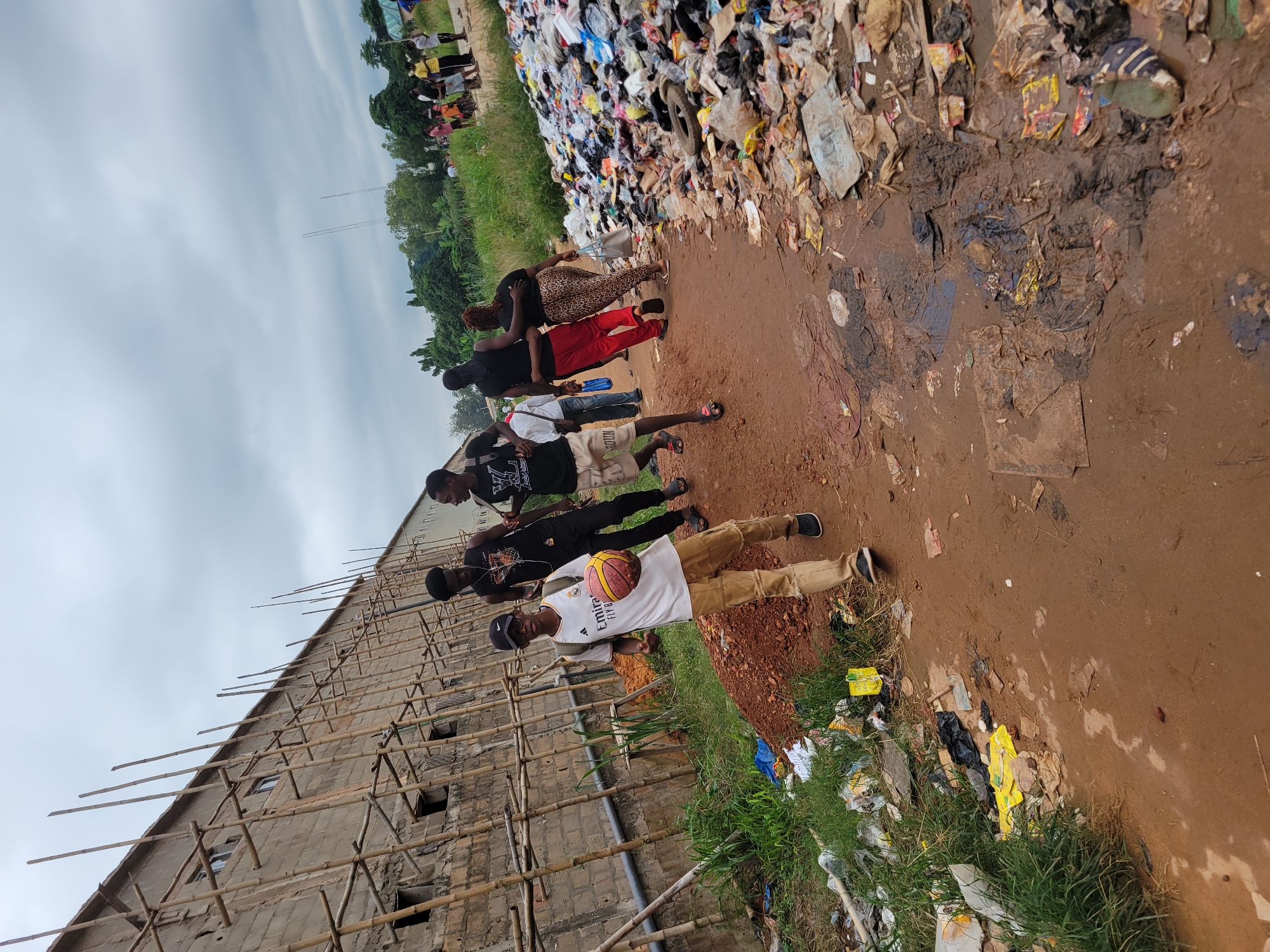 In Egbagu, Nigeria, a group of young men are seen walking along a dirt path. One of the men in the foreground, wearing a white jersey and tan pants, holds a basketball. To the right of the path is a large pile of garbage. In the background, a building under construction is visible, supported by bamboo scaffolding, and a cloudy sky looms overhead. Several other individuals are scattered in the background, some appearing to be near the construction site and others further down the path. The environment appears to be an outdoor, somewhat dilapidated area with evidence of construction and significant waste accumulation.