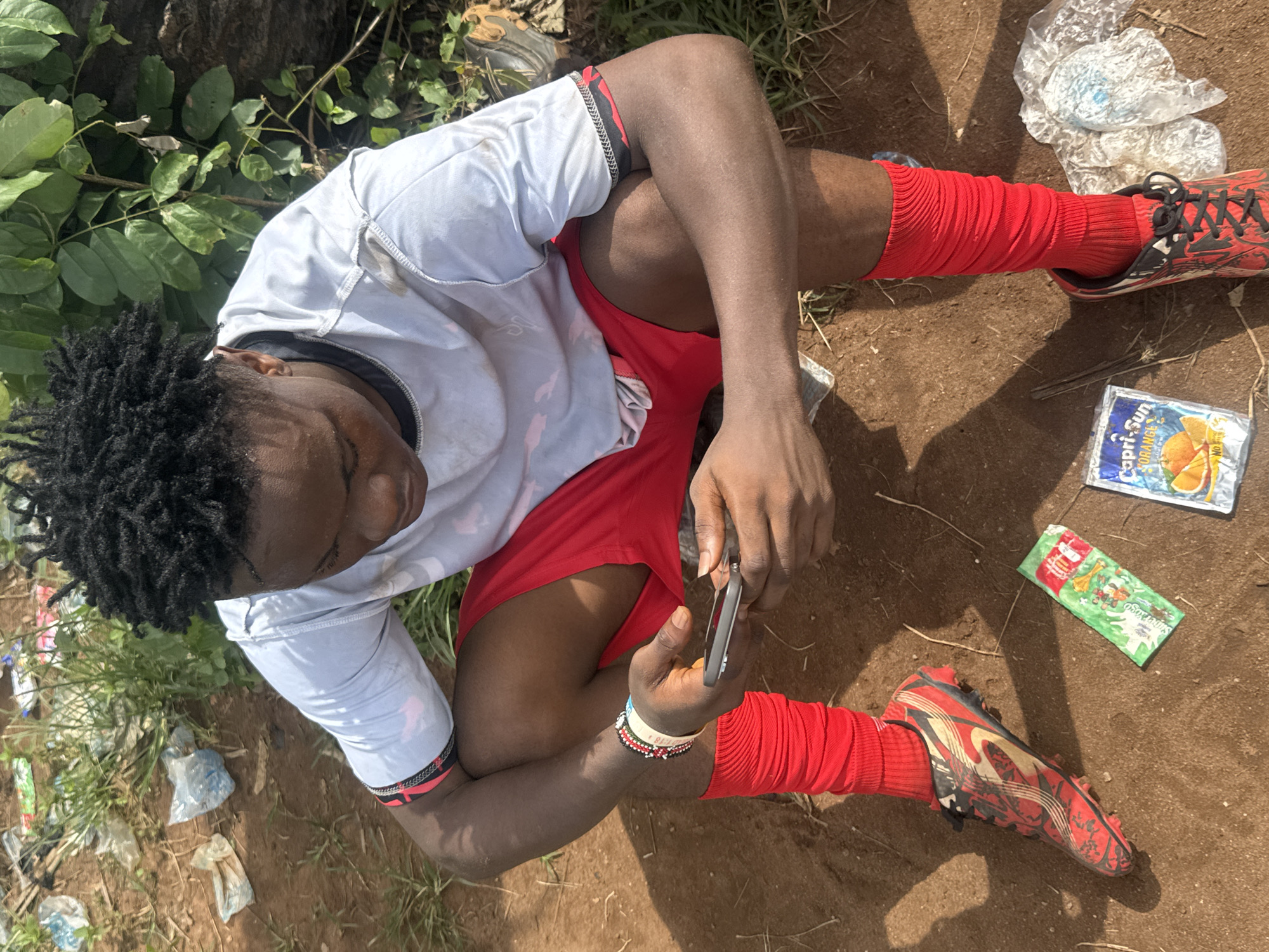 A young man is sitting on the ground, presumably outdoors, in what appears to be a partially shaded area in Ihiagwa, Nigeria. He has dark skin, short dreadlocks, and is wearing a light-colored jersey, red shorts, red socks, and red and black soccer cleats. He is looking down at a mobile phone held in his hands. Litter is scattered around him, including an empty Capri-Sun juice pouch, a green Super Yogo drink package, and plastic wrappers. The ground is reddish-brown dirt. There are green plants and a tree trunk near him.