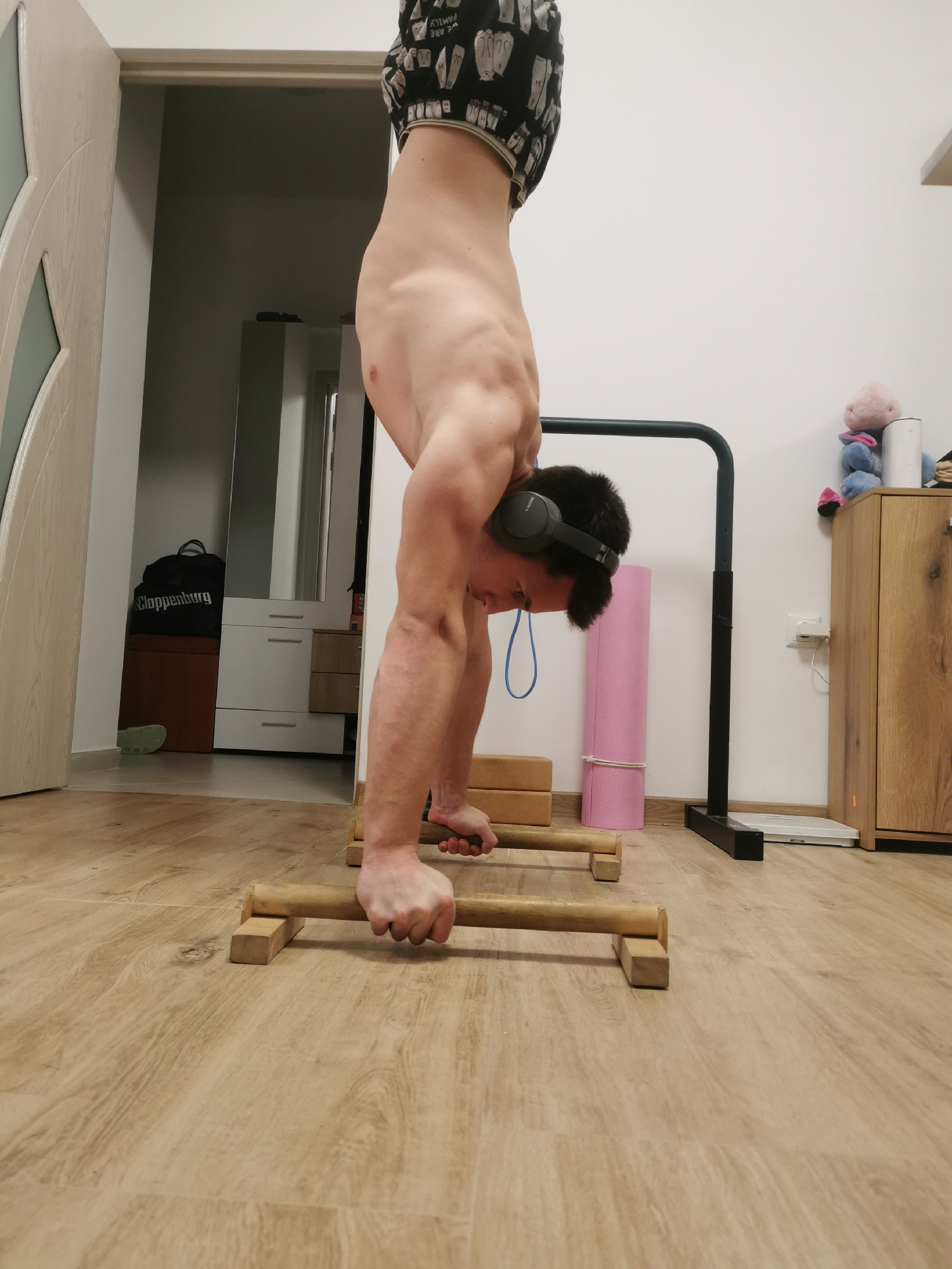 A shirtless man with black headphones is performing a handstand on wooden parallettes in an indoor setting. He is wearing patterned shorts. Behind him is a pull-up bar, a pink yoga mat, and a wooden cabinet with a stuffed animal on top. A bag labeled "Cloppenburg" is visible near a partially open door. A scale is on the floor near the pull-up bar. The location is possibly a residential space and not identifiable as Burgas, Bulgaria.