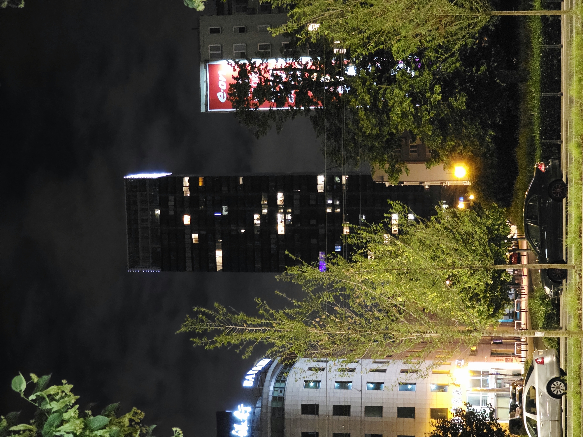 In the nighttime city scene of Warsaw, Poland, a tall, modern skyscraper with illuminated windows dominates the center of the frame. To the left, a rounded building with lit windows and blue neon signage that appears to read "BANKOWY" is visible. A large, red advertising billboard for "e-on" is mounted on a building to the right of the skyscraper. In the foreground, a silver sedan is parked on the left side of a road, and a black sedan is parked on the right. Lush green trees and foliage are scattered throughout the scene, partially obscuring the buildings. The sky is dark and cloudy.