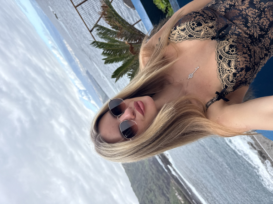 A blonde female subject, wearing hexagonal sunglasses with brown gradient lenses, is depicted in a selfie taken outdoors in Mesa Del Mar, Spain. She wears a black and gold patterned garment with lace trim, thin straps, and small black bows. A silver-colored necklace with a key-shaped pendant is visible on her chest. Her lips are painted with a pinkish tone. The background features a coastal landscape with the ocean exhibiting waves crashing against a dark, rocky shoreline on the left. Green-covered hills and mountains recede into the distance. A palm tree is visible on the right, alongside a dark metal fence and a blue wall segment. The sky overhead is predominantly cloudy.