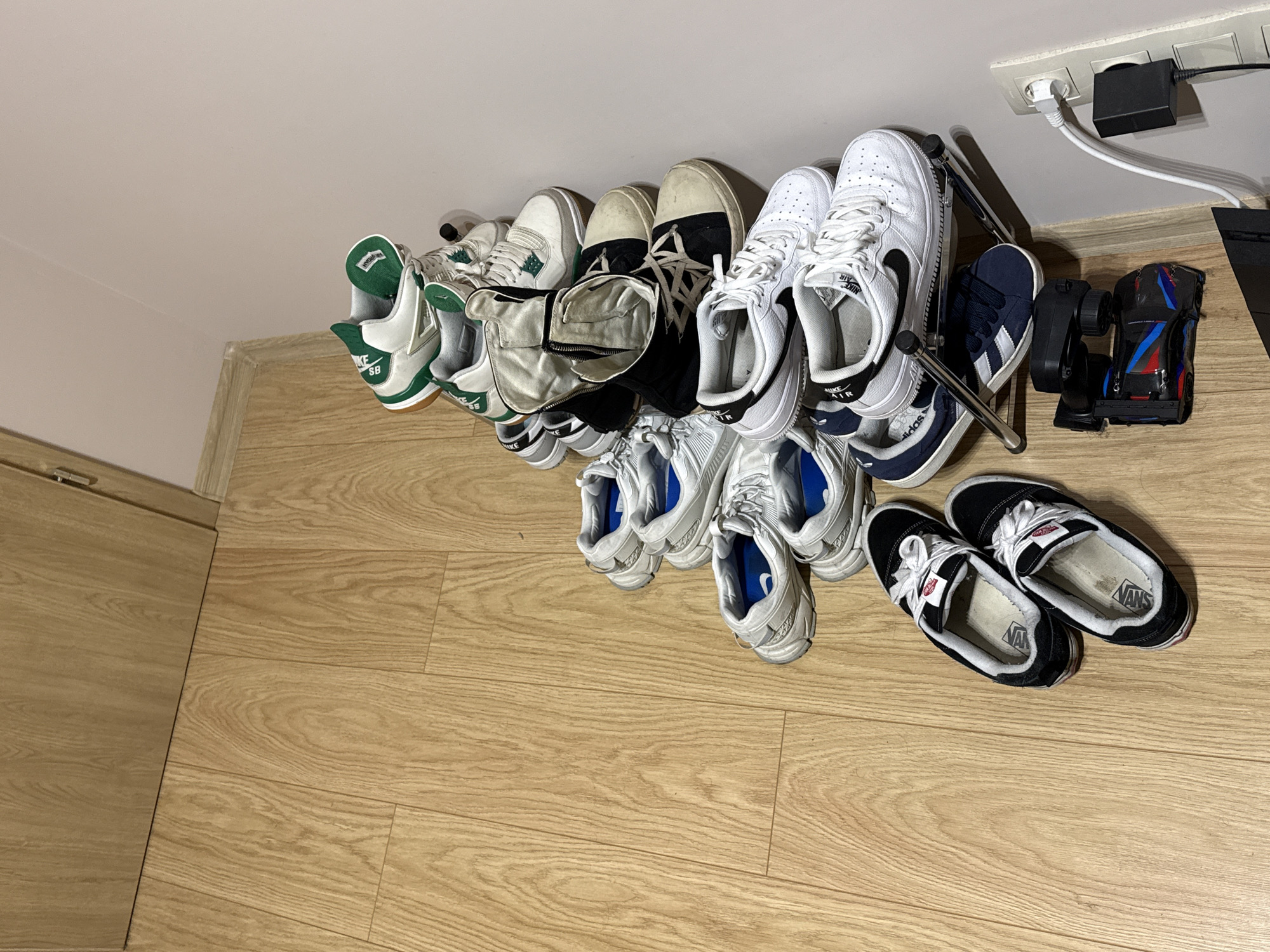 The image shows a collection of shoes arranged on a rack in a room. The floor appears to be wooden. The shoe rack is located near a wall with an electrical outlet and some electronic devices (a power adapter and what appears to be a toy car with a remote). The shoes are of various styles and brands, including Vans, Nike Air Force 1's, and Adidas sneakers. The location is inside an interior space, presumably a residence. There is no specific visual information linking the image to Sofia, Bulgaria.