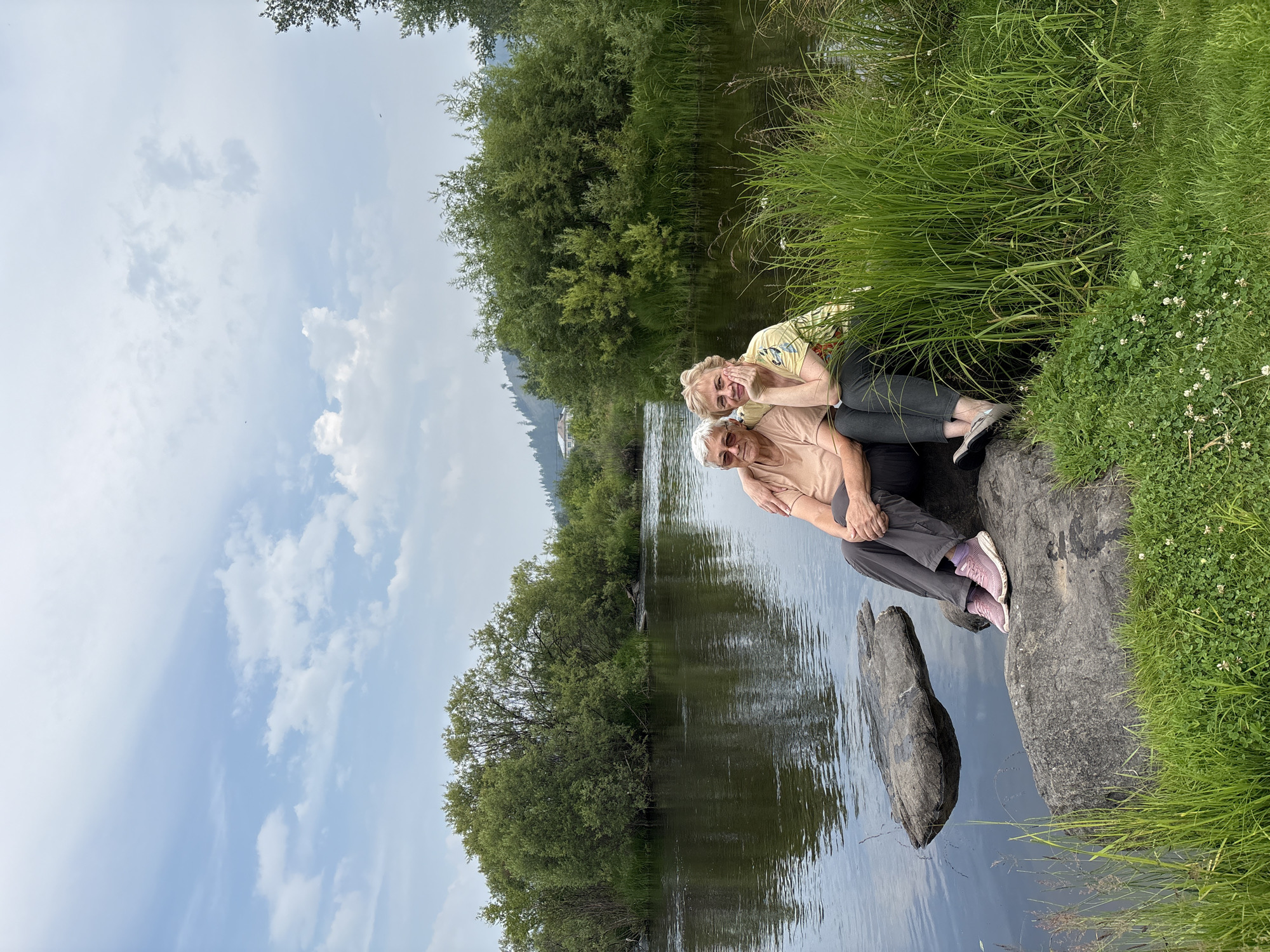 Two elderly women are seated on large rocks next to a calm river. The woman on the left, with grey hair, wears glasses, a light brown t-shirt, and grey pants. The woman on the right, with blonde hair, wears a yellow patterned t-shirt and dark pants. The woman on the left has her arm around the woman on the right. They are both looking towards the camera. The background features lush green trees and vegetation along the riverbank, with a hill and a small building visible in the distance under a cloudy sky. The river reflects the sky and surrounding greenery. The image appears to be taken in an outdoor, natural setting. The city of Невон, Russia, is not identifiable within the provided image, as it is a general landscape scene without specific landmarks or signage to confirm its location.