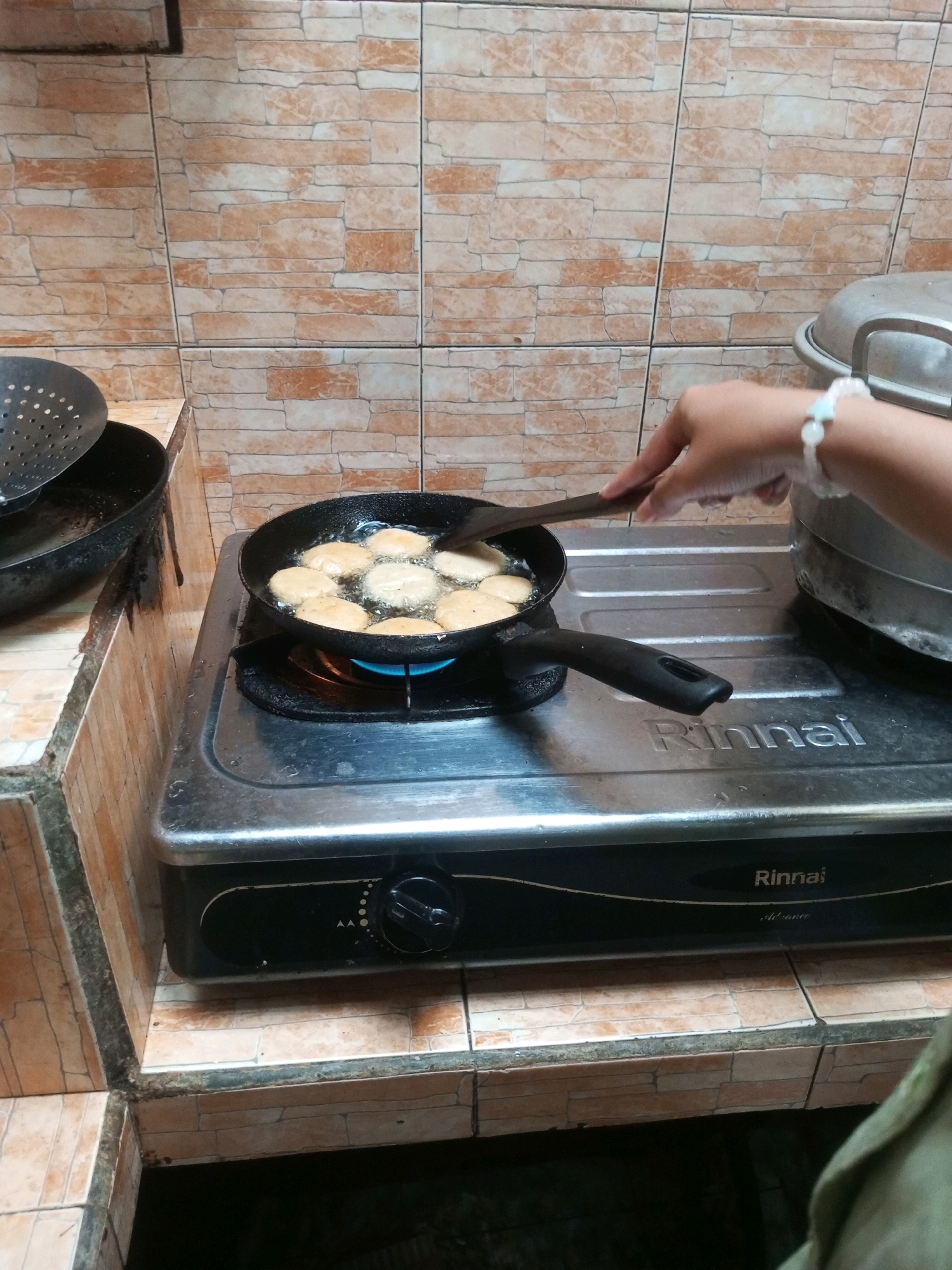 In Andongsili, Indonesia, a person is frying fritters in a black frying pan on a Rinnai gas stove. The fritters are golden brown and appear to be in hot oil. A wooden spatula is being used to turn the fritters. To the left of the stove, a metal colander rests on a black pan. The background consists of tiled walls. The scene depicts a domestic kitchen environment.