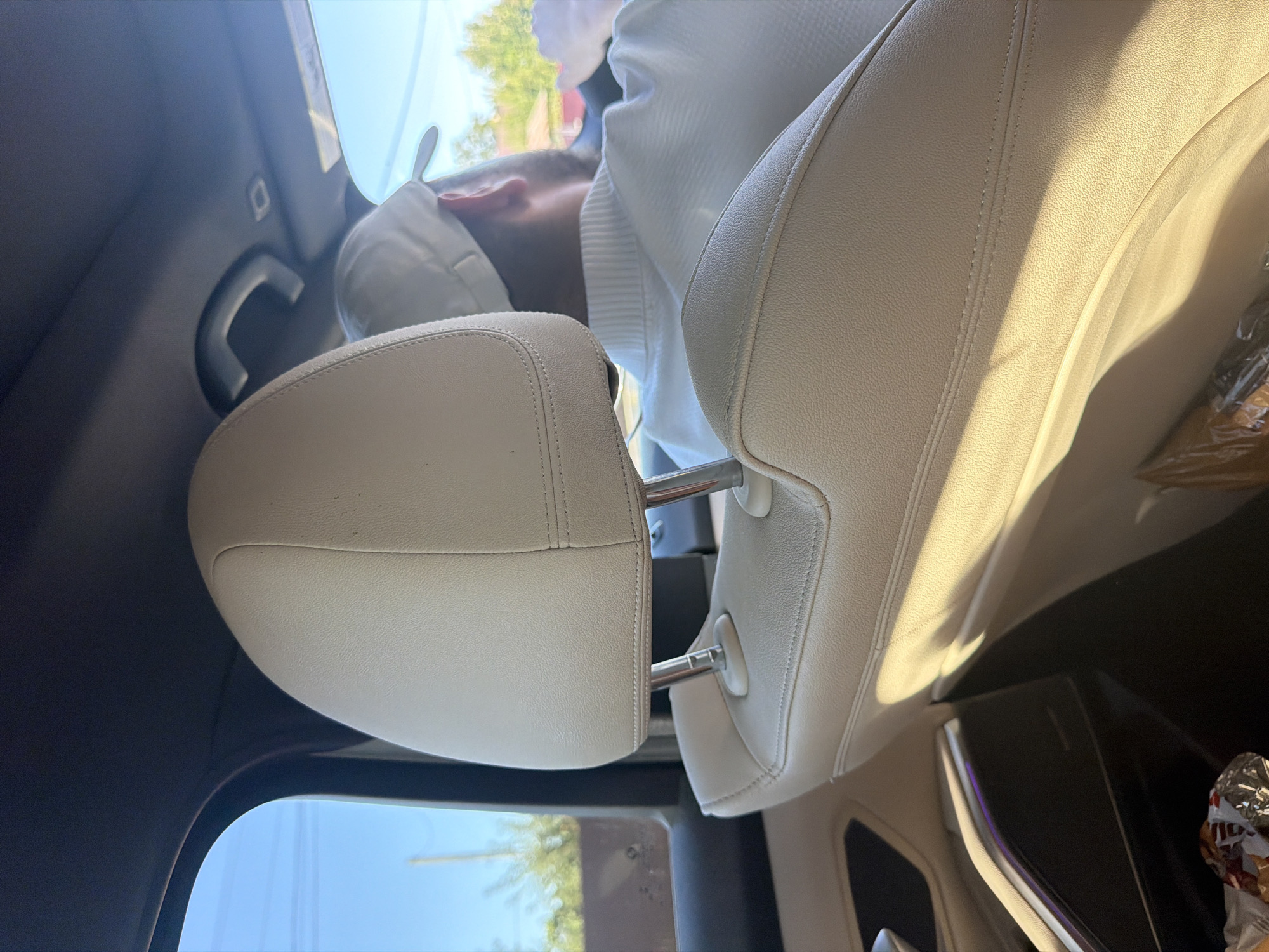 The image shows the interior of a car, viewed from the back seat, looking forward. A person wearing a white baseball cap and a white collared shirt is visible in the driver's seat, driving the car. The interior of the car features beige leather seats, with stitching detail. Sunlight streams in through the windows, casting shadows on the seats. Outside the car, through the windows, trees and a residential building are visible under a clear blue sky. The scene appears to be a person driving on a sunny day. The location is unknown and cannot be determined from the image.