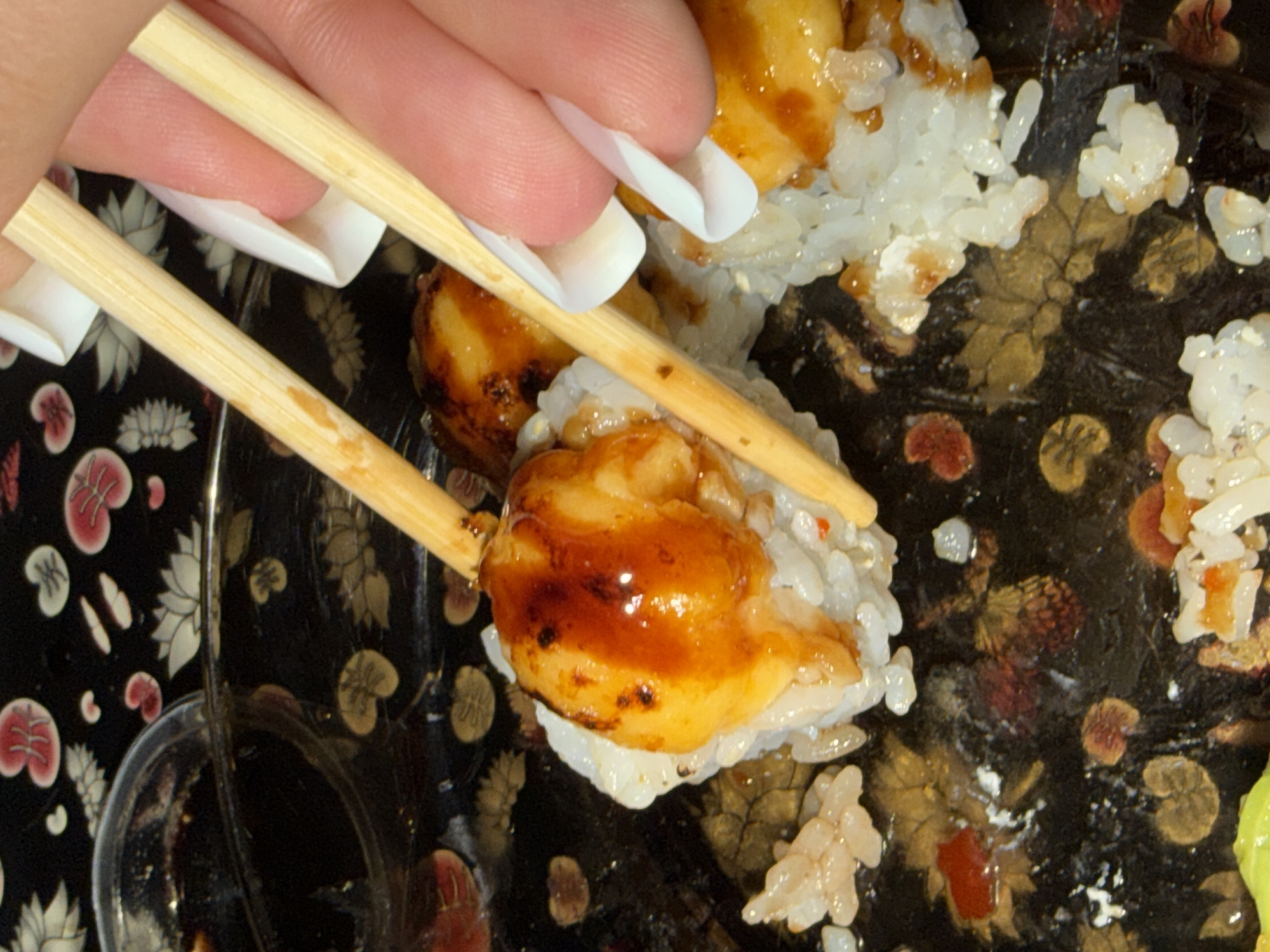 A person with white false fingernails uses chopsticks to pick up a piece of sushi, containing white rice and covered with a brown sauce. The sushi is on a black plate with a floral pattern. A small dish of dark liquid is also visible. The location is unknown and there is no evidence to suggest it is in Varna, Bulgaria.