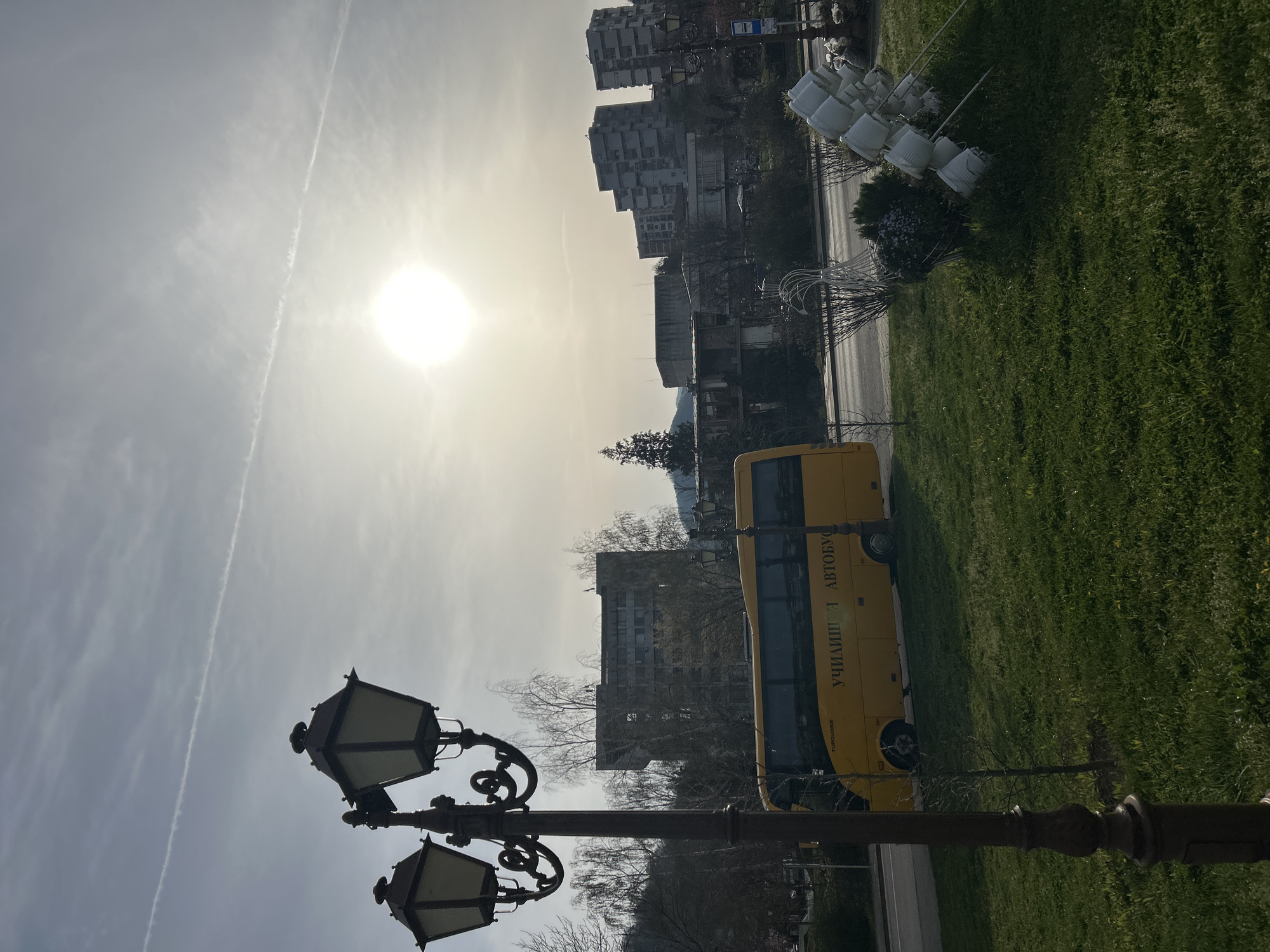 The image shows a street scene in Zlatograd, Bulgaria, taken during the day. The sun is high in the sky, with a contrail visible. In the foreground, there is an ornate black street lamp with multiple lights. To the right of the lamp post, a yellow school bus is parked on the grass. The side of the bus has text in Cyrillic, reading "УЧИЛИЩЕН АВТОБУС," meaning "School Bus." Behind the bus, there are apartment buildings. There are also several white plastic buckets arranged along the edge of the grass.