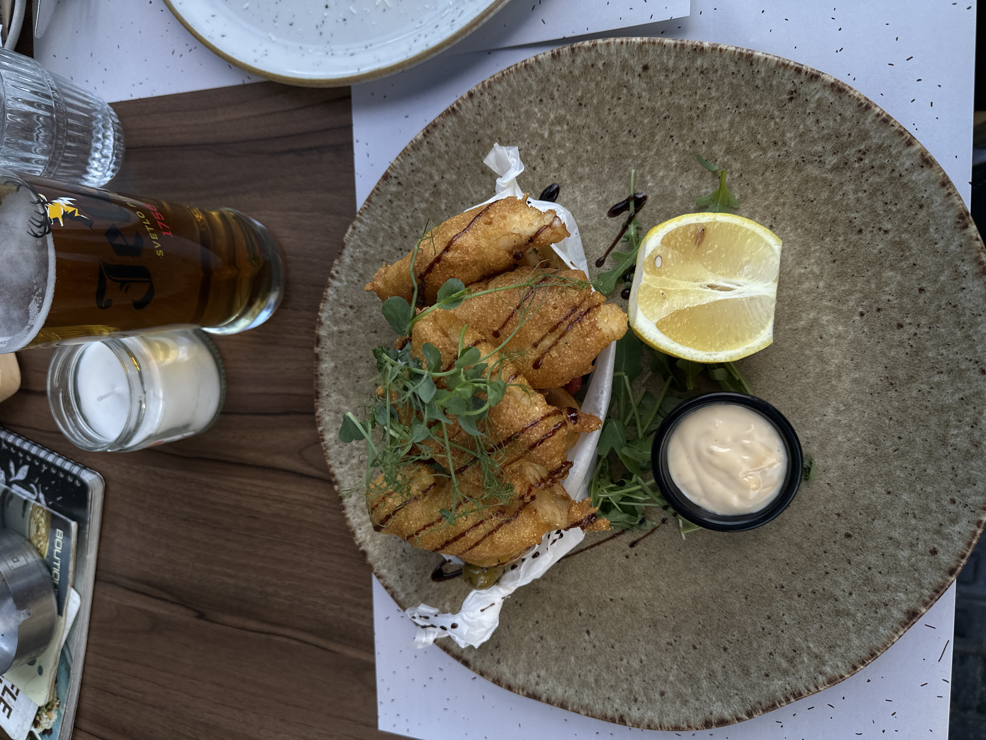 An overhead shot captures a dining scene, likely at a restaurant or cafe in Belgrade, Serbia. The primary focus is a rustic, speckled ceramic plate holding a meal. On the plate, several pieces of golden-brown, fried food, resembling fish or chicken tenders, are artfully arranged on a bed of rocket or arugula and a white paper liner. These fried items are drizzled with a dark glaze, possibly balsamic, and garnished with fresh microgreens.
Accompanying the main dish are a wedge of lemon, indicating a common accompaniment for fried food, and a small black cup filled with a creamy, white sauce, likely tartar sauce or mayonnaise. The table is made of dark wood.
To the left of the plate, a glass of light beer stands, displaying a label with a prominent stag logo. Visible text on the label includes "SVETLO PIVO" (meaning "Light Beer" in Serbian, among other Slavic languages) and the year "1756." Further to the left, a clear, textured drinking glass and a small glass jar containing a white candle are visible. In the bottom left corner, part of a black notebook with the word "BOUTIQUE" can be seen. No people are visible in the image. The lighting suggests it is daytime.