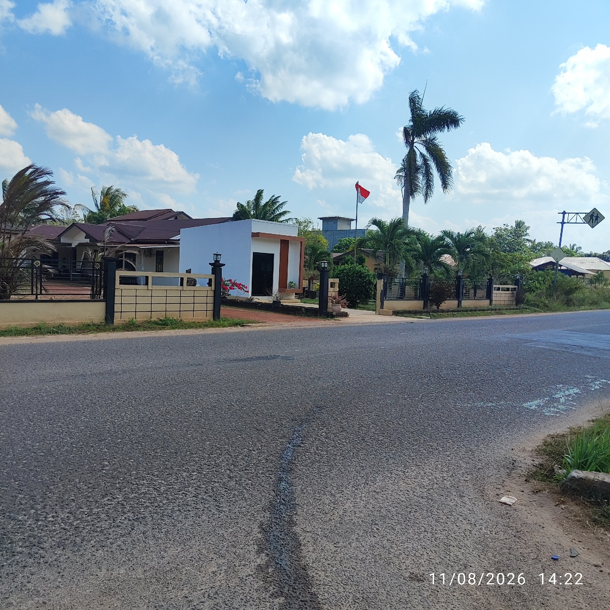 The image depicts a residential property in the city of Pembaharuan, Indonesia, on a sunny day with a partly cloudy sky. A paved road runs in the foreground, bordering a property enclosed by a fence. The fence has a combination of dark metal bars and a lighter colored concrete or stone base.

Within the property, several buildings are visible. A modern, white building with a dark doorway and wooden accents is prominent. Adjacent to this is a larger, older-style house with a reddish-brown, gabled roof. Both structures appear to be well-maintained. Tropical palm trees and other dense green foliage surround the buildings, suggesting a warm climate.

In front of the white building, a tall flagpole stands, flying a red and white flag, which is the national flag of Indonesia. A school crossing sign, with two stick figures walking, is visible on a blue pole to the right of the frame, indicating the proximity of a school or a pedestrian crossing area. The scene suggests a peaceful, everyday setting in a suburban or semi-rural area of Pembaharuan. A timestamp in the lower right corner reads "11/08/2026 14:22".
