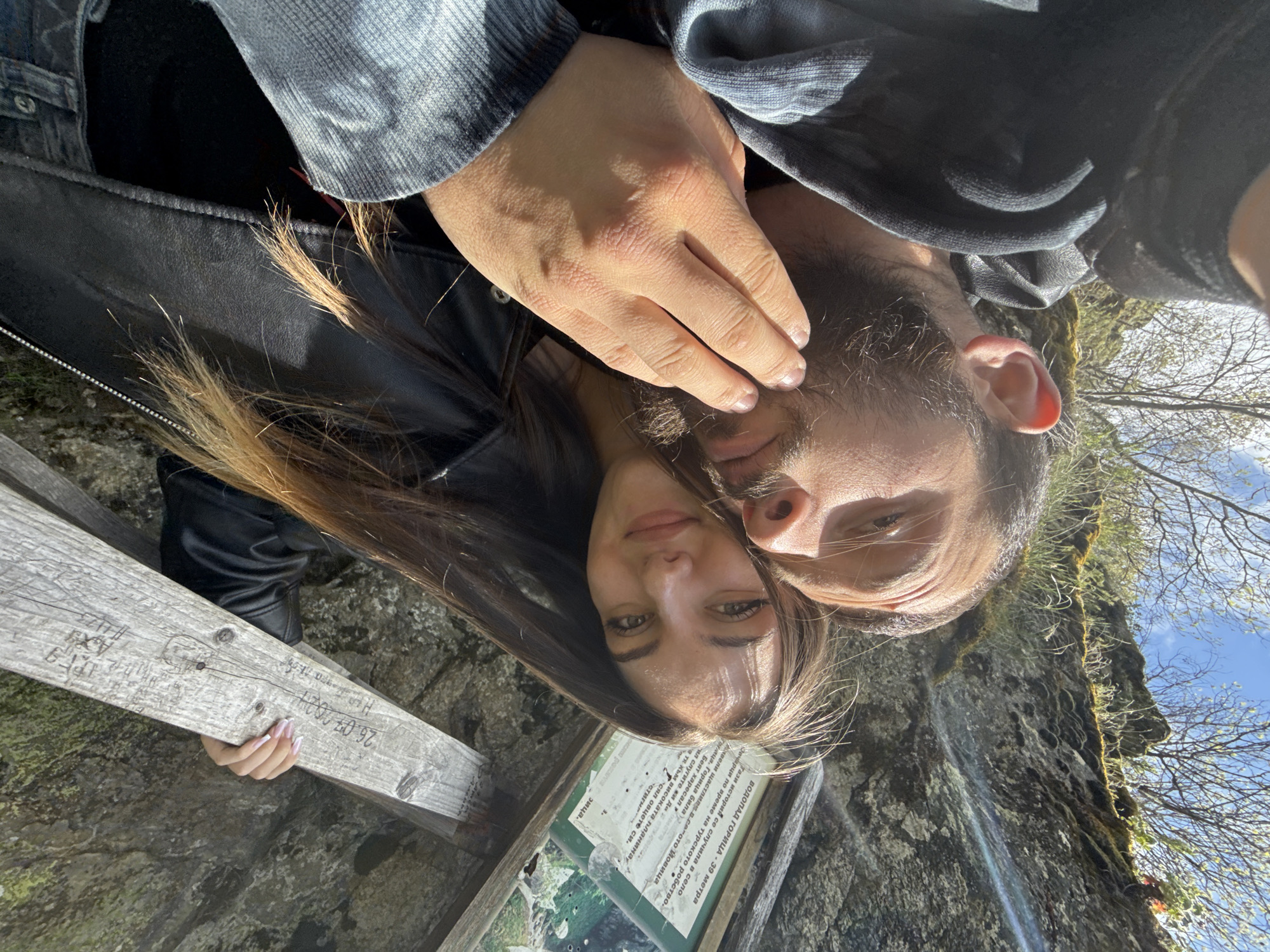 A man and woman are posing for a selfie in front of a waterfall. The woman has long brown hair and is wearing a black leather jacket. The man has a beard and is wearing a dark hoodie. Behind them is a rocky cliff with moss and trees. There is a wooden sign with carved writing, and a green information board. The OCR text on the information board contains information about the Goritsa Waterfall near Ovchartsi, Bulgaria.