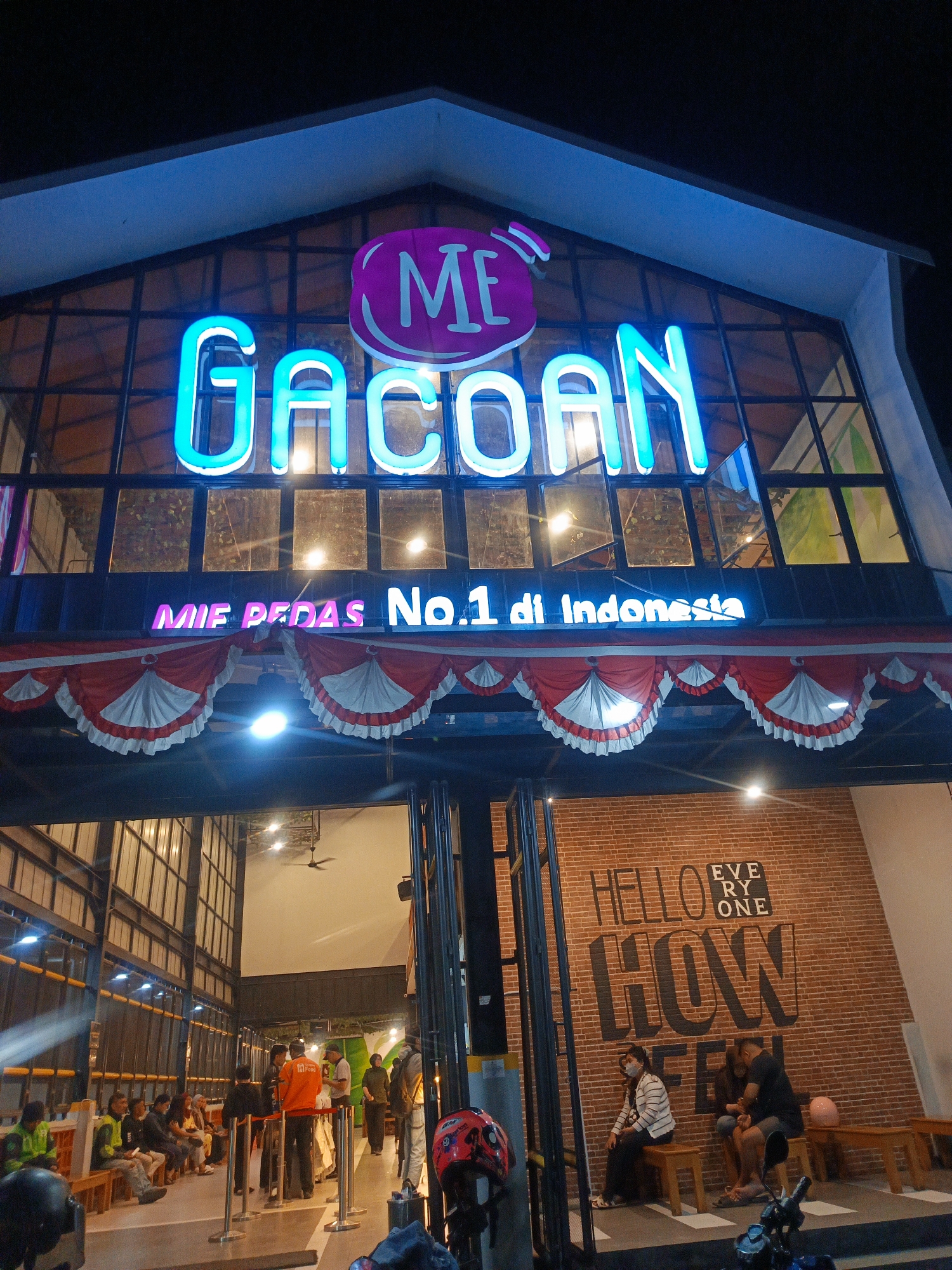 The image depicts the exterior of a restaurant at night, likely in Gorontalo, Indonesia, identified by the neon sign that reads "ME GACOAN MIE PEDAS No.1 di Indonesia". The building has a modern facade with large glass windows and an angular roof structure. Red and white decorative fabric banners hang above the entrance. Inside, people are seen sitting and standing, with some appearing to be staff members. To the right, a brick wall features large lettering that reads "HELLO EVERYONE HOW FEEL". A motorcycle is parked in the foreground on the right side. The overall ambiance is that of a popular eatery with bright neon signage and a bustling atmosphere.