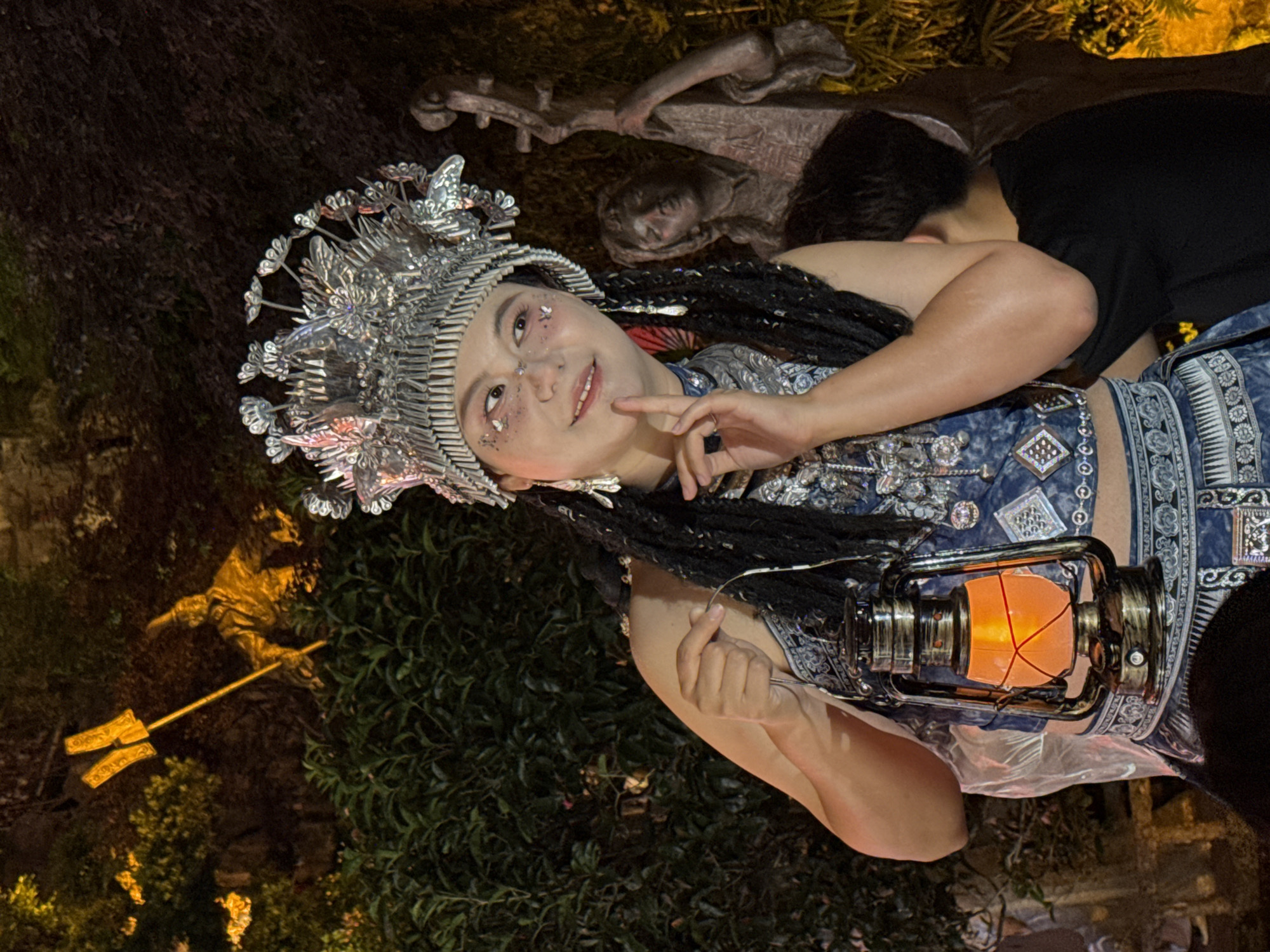 A woman wearing a decorative silver headdress and a traditional blue and silver outfit holds a lit lantern in an outdoor setting. She is turned slightly to the right, looking up and to her left, with a finger pointing to her cheek. A person wearing a black shirt is partially visible behind her. In the background, there are various decorative elements, including sculptures and a statue with a trident. The setting appears to be Yuzhong District, China, based on the visual elements that suggest a cultural performance or festival.