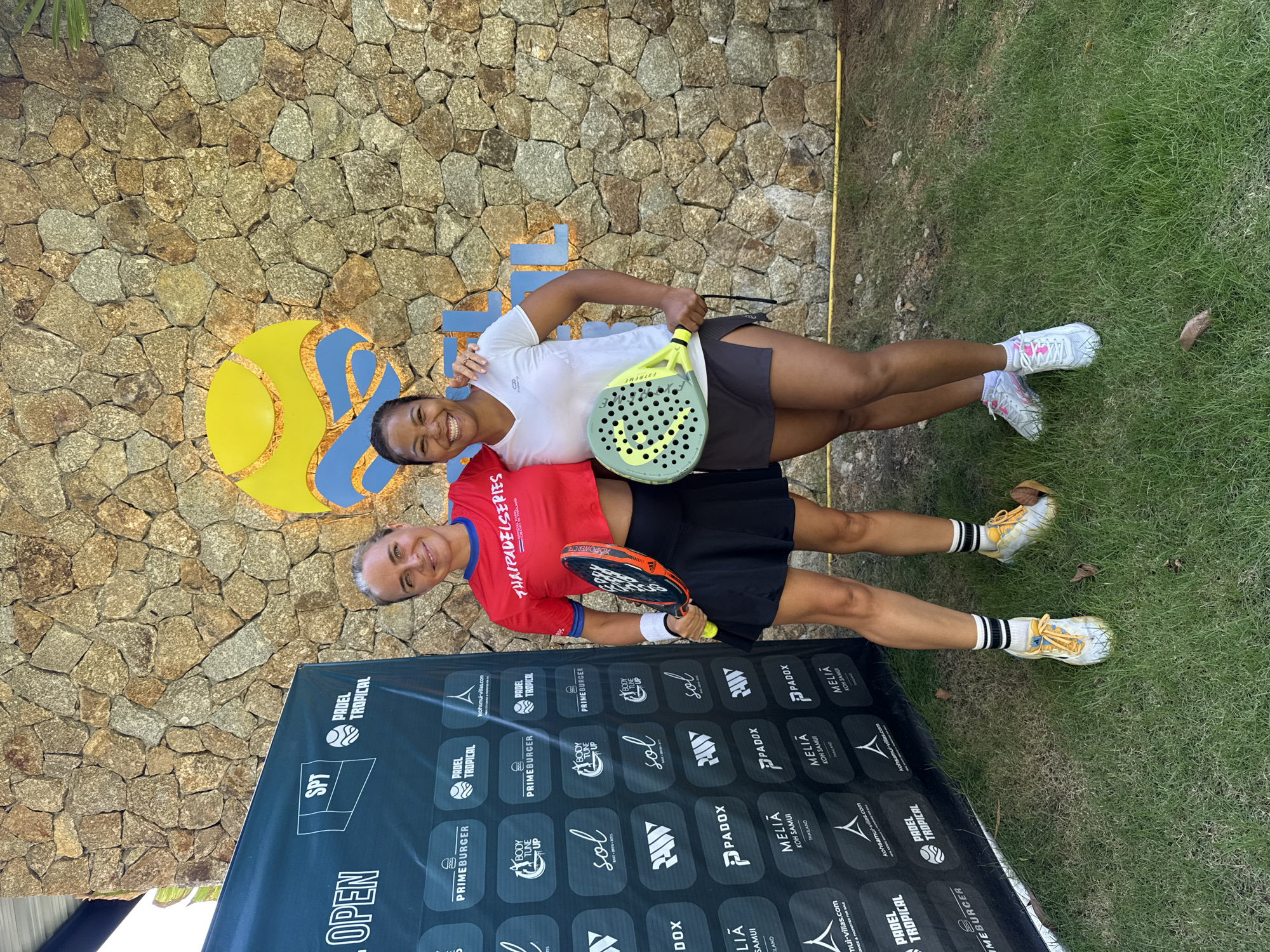 Two women, posed with padel rackets, stand in front of a stone wall backdrop in บ้านบางรักษ์, Thailand. The woman on the left, with blonde hair, wears a red cropped t-shirt with "THATPADELSERIES" printed on it, a black pleated skirt, white socks with black stripes, and yellow and white athletic shoes. She holds a black and orange padel racket. The woman on the right, with dark hair, wears a white t-shirt, a dark grey skort, white athletic socks, and white athletic shoes with pink accents. She holds a light green and yellow padel racket. A black banner with various logos, including "SPT" and "PADEL TROPICAL," is positioned to the left of the women. A large, colorful logo featuring a yellow circle and blue wavy lines is behind and above them. The ground in front of the subjects is covered in green grass.