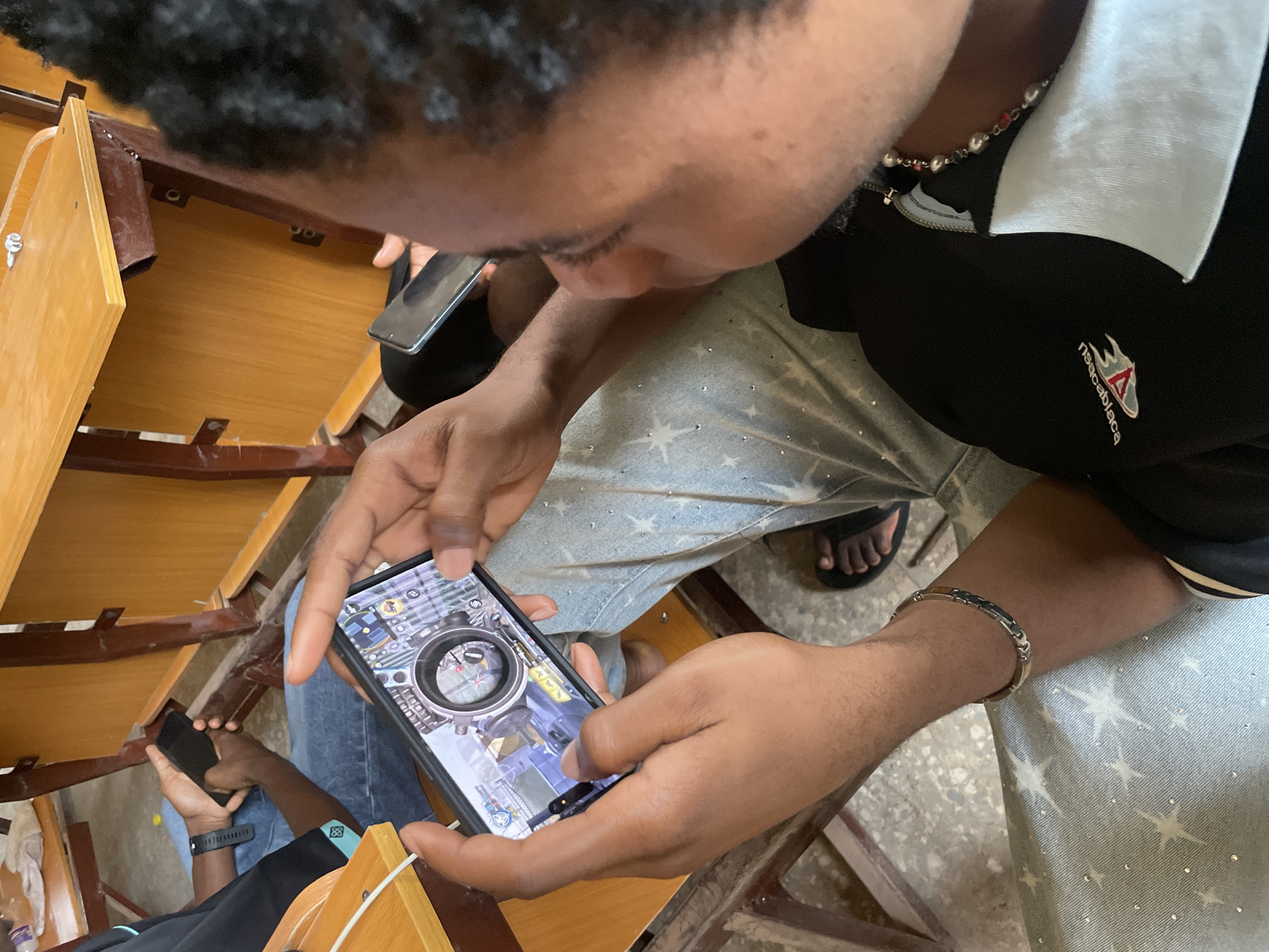The image shows a close-up, overhead view of two individuals sitting at school desks in what appears to be a classroom setting in Ihiagwa, Nigeria. The primary focus is on a person holding a smartphone in landscape orientation, actively engaged in playing a mobile game. The game displayed on the screen appears to be a first-person shooter or action game, with a scope and in-game elements visible. The person's hands are positioned to control the game, with thumbs on the touchscreen. Another individual in the background is also holding a smartphone, though their screen is not as clearly visible. Both individuals are wearing casual clothing. The environment suggests a functional, possibly modest, educational facility with wooden desks and chairs. The lighting appears to be ambient indoor lighting.