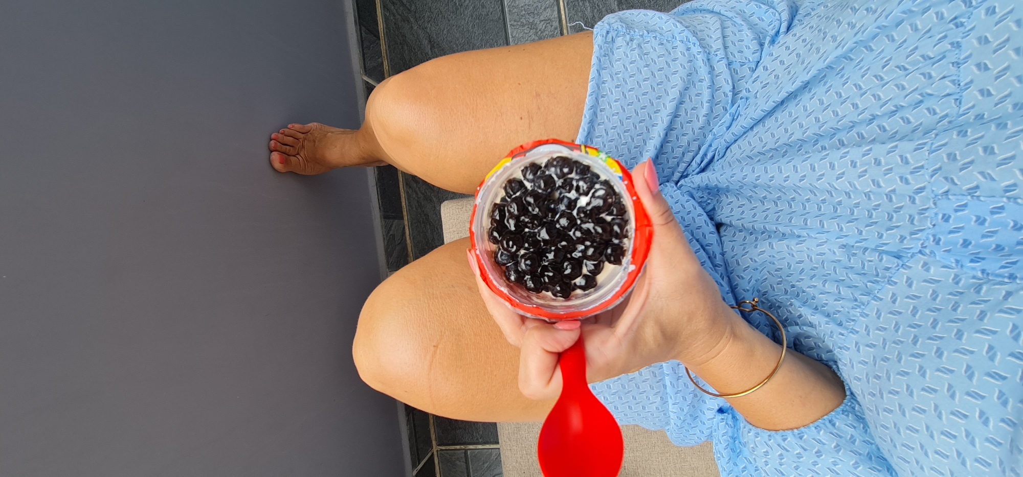 This high-angle, first-person perspective image captures a person sitting in what appears to be an outdoor or semi-outdoor setting in Da Nang, Vietnam. The primary focus is on the person's hands and lap as they hold a cup of food.
The person is wearing a light blue dress or top with a distinctive pattern of small, repeating light blue and white geometric shapes. Their bare legs, showing tanned skin, are extended forward. The right foot, adorned with red nail polish on the toes, rests on a smooth, dark gray surface. A hand, with pink nail polish, holds a clear cup that has a vibrant red rim and possibly other colorful details on its exterior. The cup is filled with numerous dark, glossy, spherical objects, resembling tapioca pearls or boba, suspended in a creamy white substance, likely a yogurt or dessert base. A bright red spoon is held in the right hand, positioned above the cup as if about to scoop its contents. A gold bangle bracelet is visible on the person's right wrist.
The background consists of the dark gray surface extending to the left and foreground. To the right of the person's legs, dark gray tiled flooring with lighter grout lines can be seen, suggesting a patio or balcony area. The lighting is bright and even, indicating a daytime scene. The image conveys a sense of relaxed enjoyment. There is no visible text.