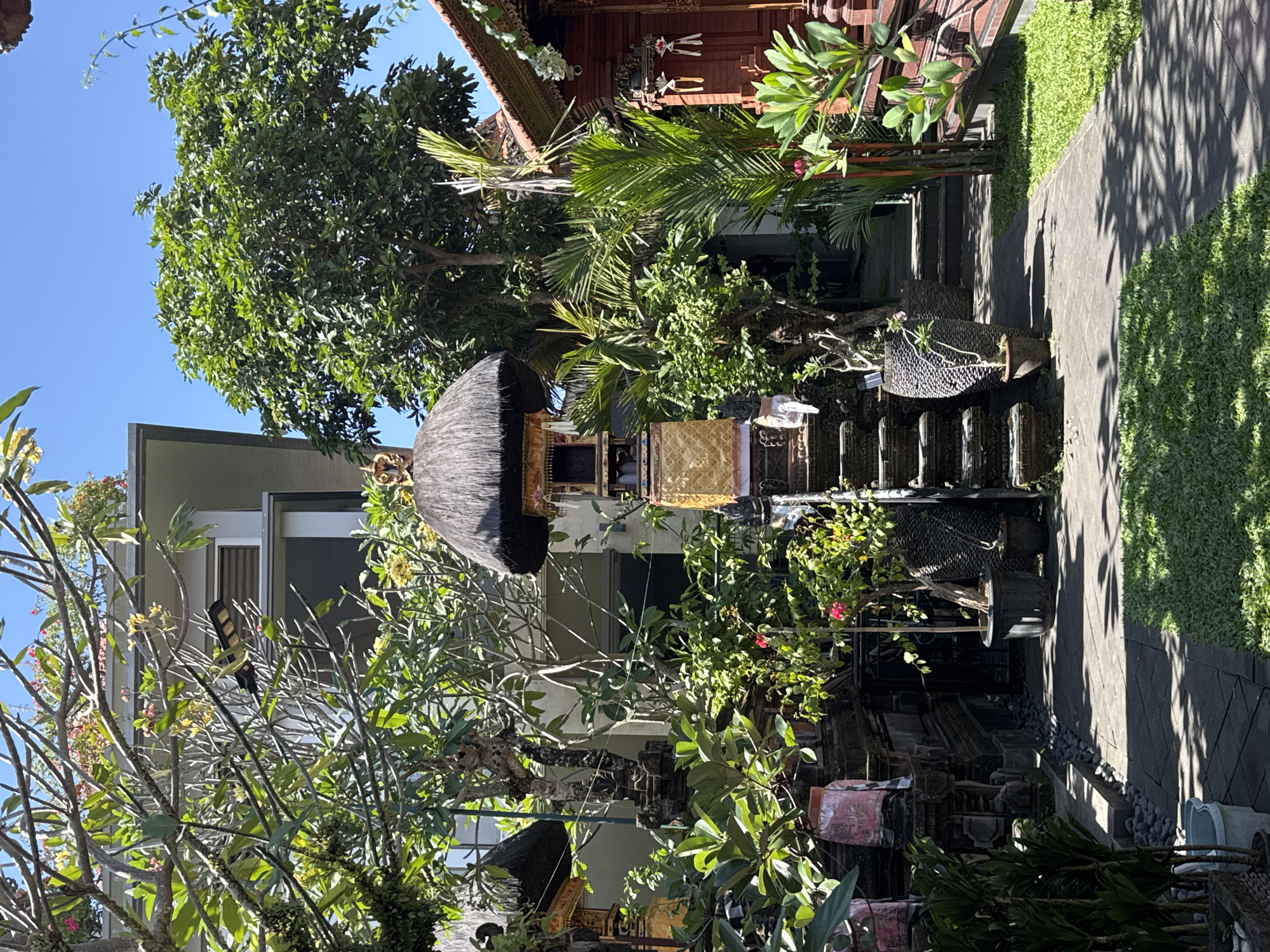 This image depicts an outdoor scene in what appears to be a courtyard or garden area, likely in North Kuta, Indonesia. The setting is lush and tropical, with abundant greenery from trees and plants. In the foreground, a small structure with a thatched roof and ornate golden detailing is visible, suggesting a decorative or spiritual element. Steps, paved with dark stones, lead upwards, bordered by a well-maintained green lawn on the right. Shadows from overhead foliage are cast across the paved path, indicating it is daytime, likely with bright sunlight.
The overall impression is one of tranquility and cultural richness. The architecture and decorative elements hint at a traditional Indonesian or Balinese style. There are no people visible in the image, but the presence of what might be offerings or decorative items near the thatched structure suggests it is a place of some significance. The scene is bathed in sunlight, creating a warm and inviting atmosphere. The focus is on the natural elements intertwined with man-made structures, creating a harmonious composition. The image captures a serene moment in a seemingly well-kept outdoor space.