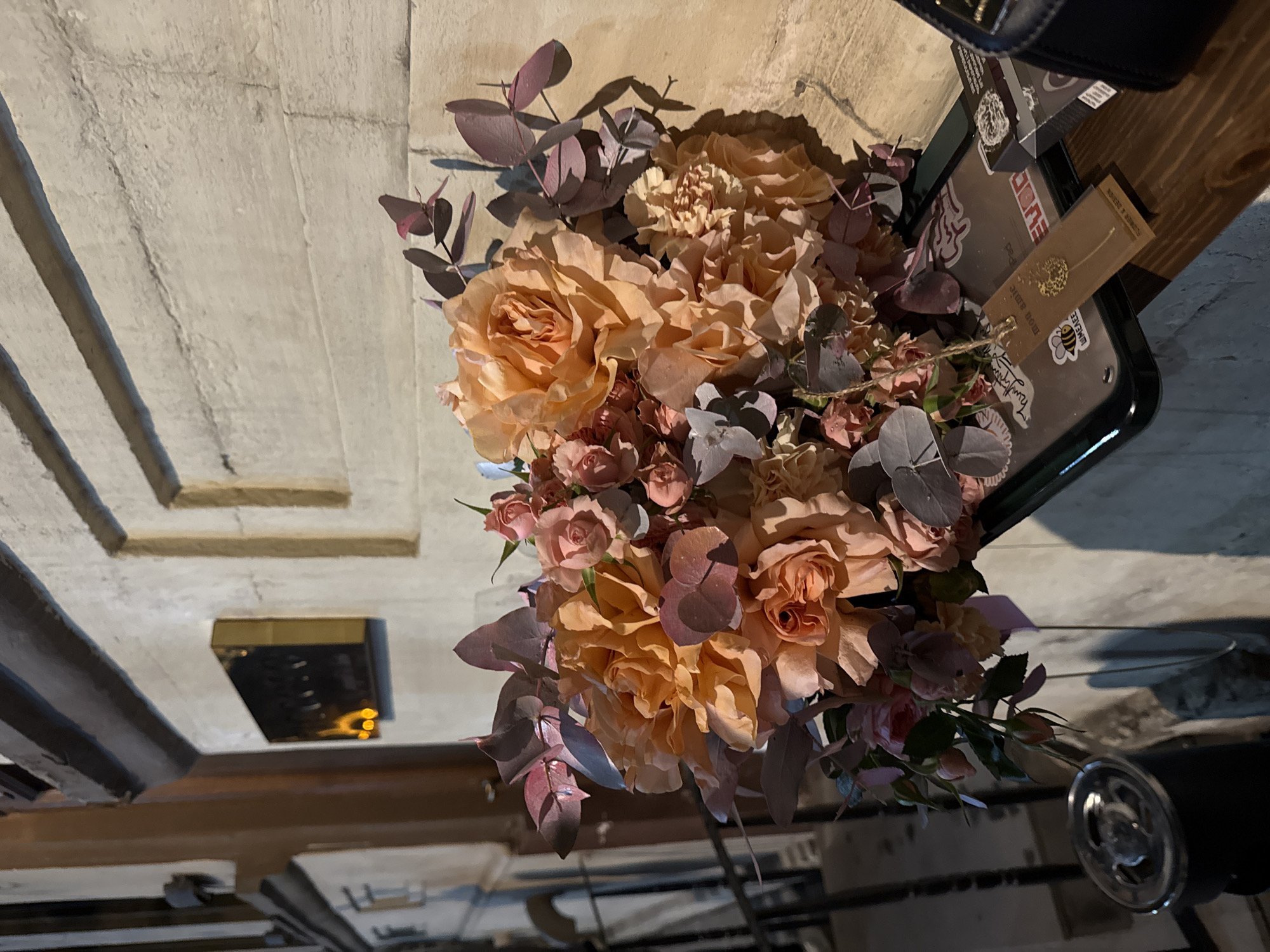 A bouquet of peach and pink flowers with purplish-grey foliage is positioned on a table, partially obscuring a dark tray with stickers and a small tag. The tray seems to be sitting on or near a purse. The setting appears to be indoors, with a decorative panelled wall in the background. The location is suggested to be in Yerevan, Armenia, but the image itself does not offer specific geographical information to confirm this detail.