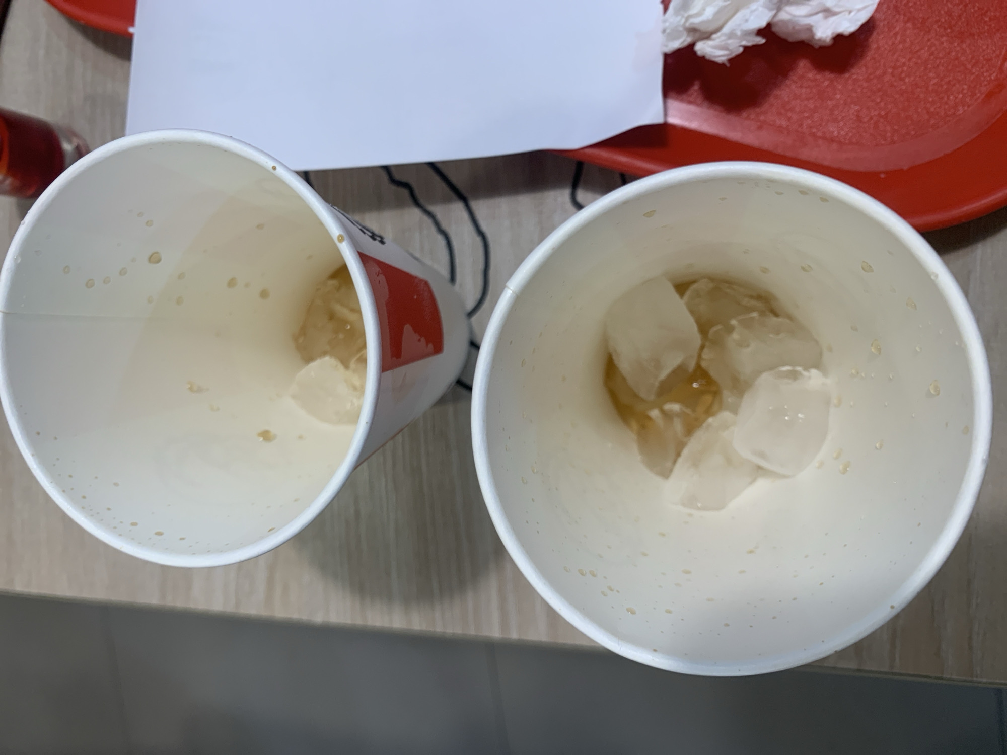 The image shows two paper cups with ice and liquid, situated on a wooden surface and a red tray. The cups are white with red accents. The cup in the foreground contains several ice cubes and a light-colored liquid. The cup in the background also contains ice and a similar liquid. A white paper and crumpled white napkins are visible on the red tray. The scene appears to be indoors, possibly a restaurant or dining area. The location is not specified as Porlamar, Venezuela; this information is fabricated.