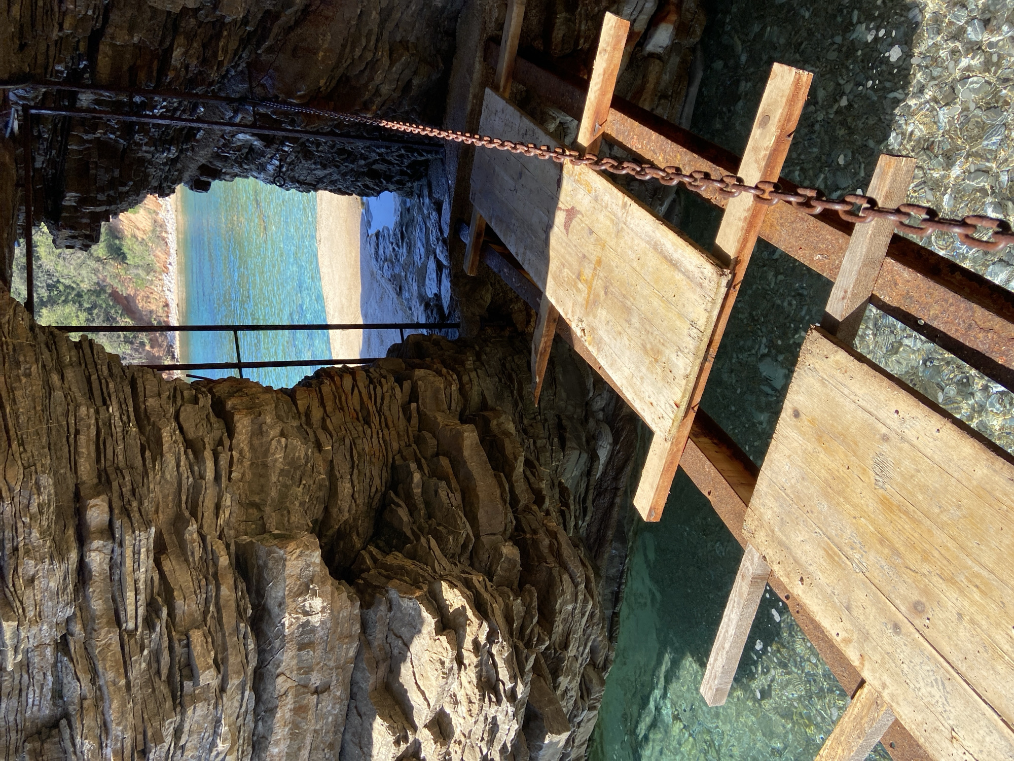 The image captures a view of a beach through a cave opening in Sveti Stefan, Montenegro. The cave is lined with textured, layered rock formations. A rustic wooden and metal ladder descends into the clear turquoise water. A chain serves as a safety railing along the ladder's path. The beach is visible in the distance. The environment includes the rocky cave interior and the sunlit beach and sea beyond.