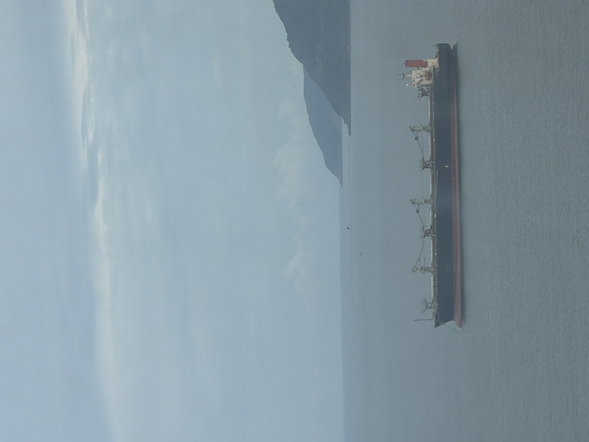 An elevated, wide shot captures a vast expanse of overcast ocean meeting a distant, misty coastline. In the foreground and mid-ground, a large, dark cargo ship with a red waterline is the primary subject. The vessel has several cranes visible on its deck. The sea is calm, with subtle ripples. In the background, two large, dark, mountainous landmasses are visible, their forms softened by the atmospheric haze. The sky is a uniform light gray, indicating cloudy or foggy conditions. No people are visible in the frame. The scene appears to be set in a coastal area, potentially near a port or along a shipping route. Given the description of a cargo ship and mountainous coastline, it is plausible that this image was taken in the vicinity of Hải Vân Ward, Vietnam, which is known for its scenic coastal mountains and proximity to the sea.