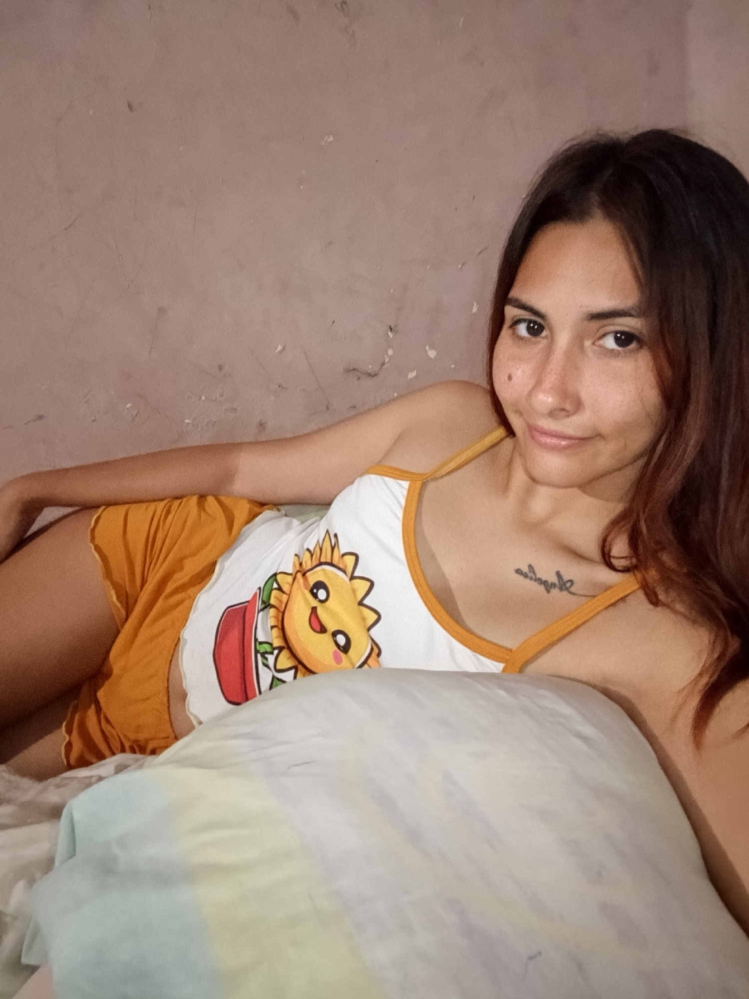The image shows a young woman with long, dark brown hair lying on a bed. She is wearing a white tank top with a cartoon sunflower design and orange shorts. She is looking at the camera with a slight smile. A pillow is in the foreground, partially obscuring her body. The background appears to be a textured wall, possibly in a bedroom. There is no specific indication of the location being Juan Griego, Venezuela, or any other city. The scene depicts a casual, indoor setting.