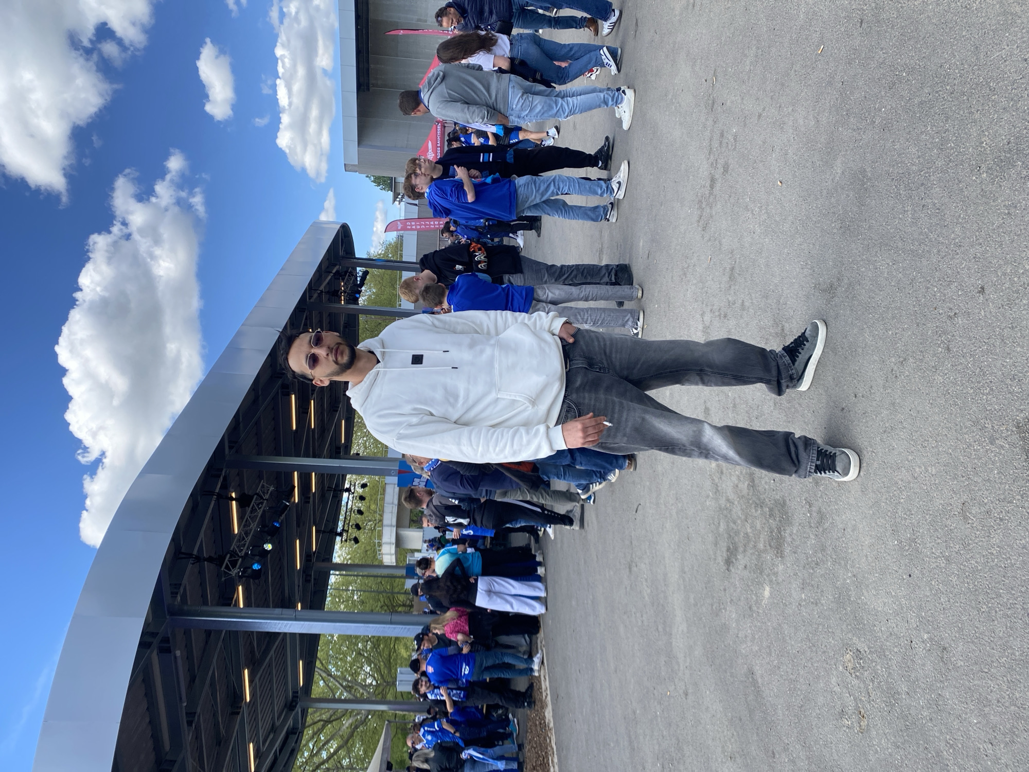 The image shows a man standing in front of a crowd of people in Strasbourg, France. The man is wearing a white hoodie, gray jeans, and sunglasses. He is holding a cigarette. The crowd is mostly wearing blue clothing, and they are standing in a line. The background includes a large metal structure with lights and a blue sky with white clouds. The surface is pavement.