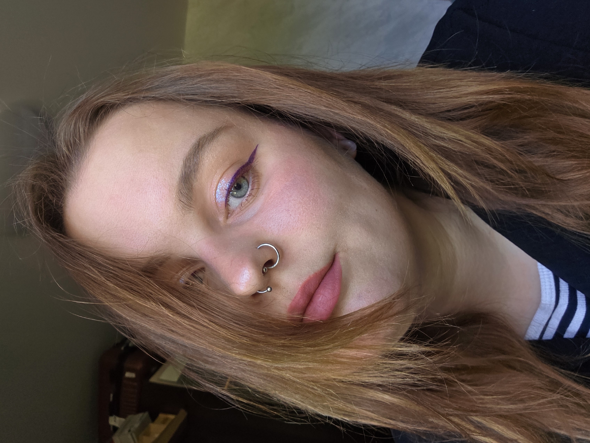 A close-up shot features a person with long, light auburn hair partially obscuring their face. The individual has fair skin, light-colored eyes, and noticeable makeup. Their left eye is adorned with a line of purple eyeliner extending outwards. They also appear to have iridescent eyeshadow and light pink blush. A small silver hoop and a small silver stud are visible in their left nostril. Their lips are painted a matte mauve color. The person appears to be wearing a black shirt with a white striped collar. The background is indistinct, featuring muted tones, possibly from an interior space.