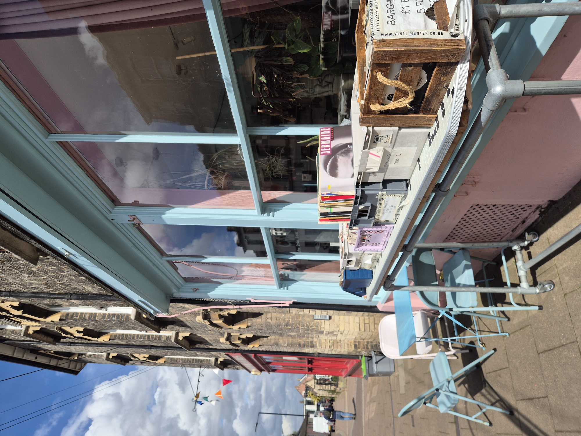 Here is a detailed description of the image:
**Scene:** An outdoor shot of what appears to be a storefront in a West Suffolk, United Kingdom setting. The perspective is somewhat tilted, with the building facade positioned along the left side of the frame and the sidewalk along the right.
**Architecture and Storefront:** The building is a mix of brick and stucco, with sections painted in pastel colors (pink and light blue). There's a large window, reflecting the sky and clouds, under which is a display shelf supported by a metal frame. This shelf is populated with various items, including a wooden box containing what appear to be vinyl records and related promotional material.
**Furniture and Sidewalk:** Several light blue folding chairs are arranged on the sidewalk, suggesting an outdoor seating area. The sidewalk is plain and grey.
**Objects and Details:** The storefront window displays plants and reflections of the surrounding environment. The shelf contains a mix of records, promotional papers, and a wooden crate filled with records and ephemera. Pennants are visible in the background.
**People:** A lone individual is captured in the background walking down the sidewalk.
**Environment:** The scene is taken on a bright day with a partly cloudy sky. The overall atmosphere is typical of a small, quaint business district.