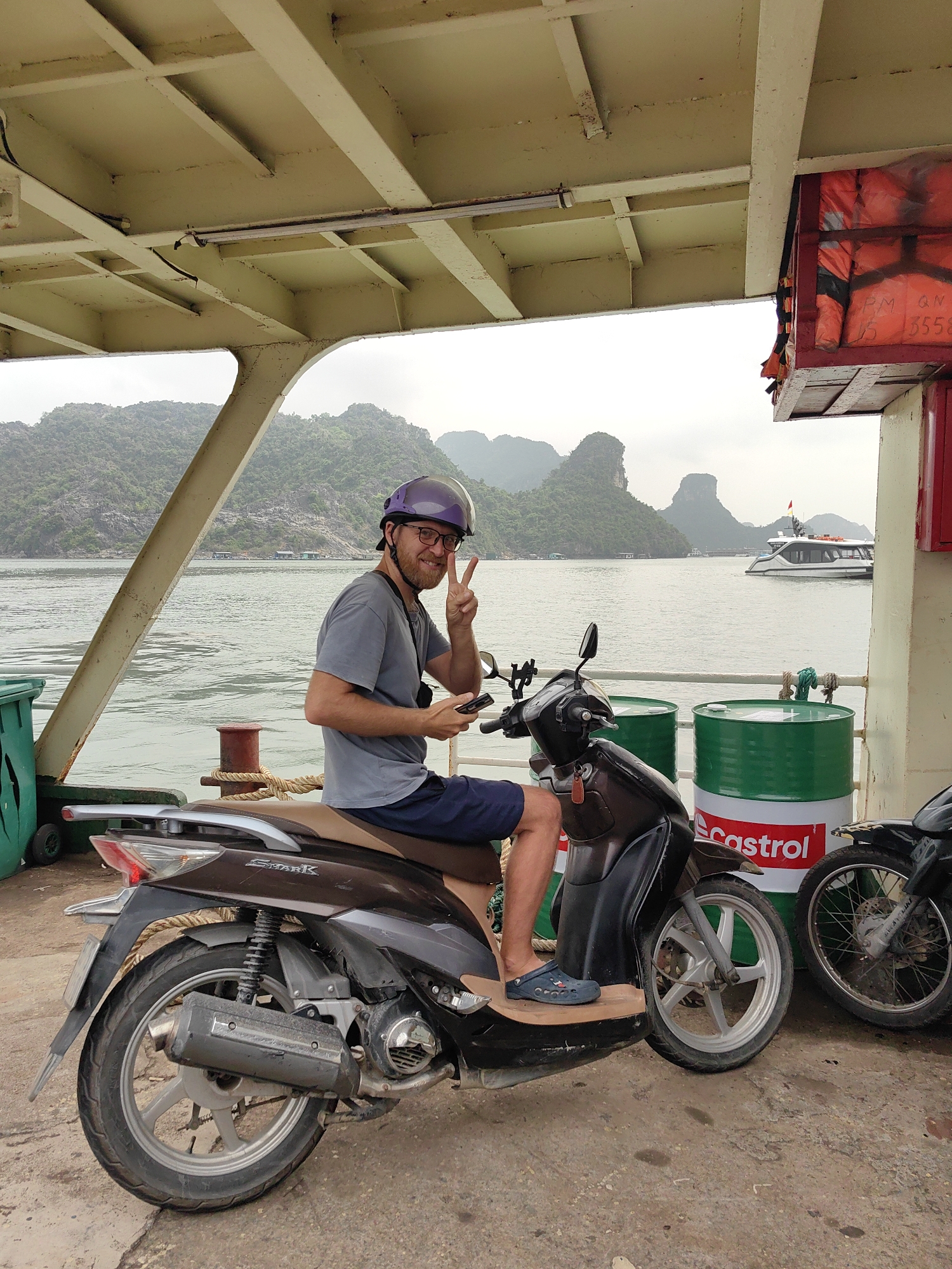 A man sits on a brown scooter on what appears to be a ferry or barge. He wears a grey t-shirt, blue shorts, and a purple helmet. He is holding a phone and making a peace sign with his left hand. Behind him are green drums with the Castrol logo. Other motorcycles are parked nearby. In the background, there's water and a landscape of green, mountainous islands. A white boat is also visible in the water. A metal awning covers the ferry deck above. A rack of orange life preservers is also visible. The scene appears to be in or near the city of Hải Phòng, Vietnam, based on the landscape and maritime setting.