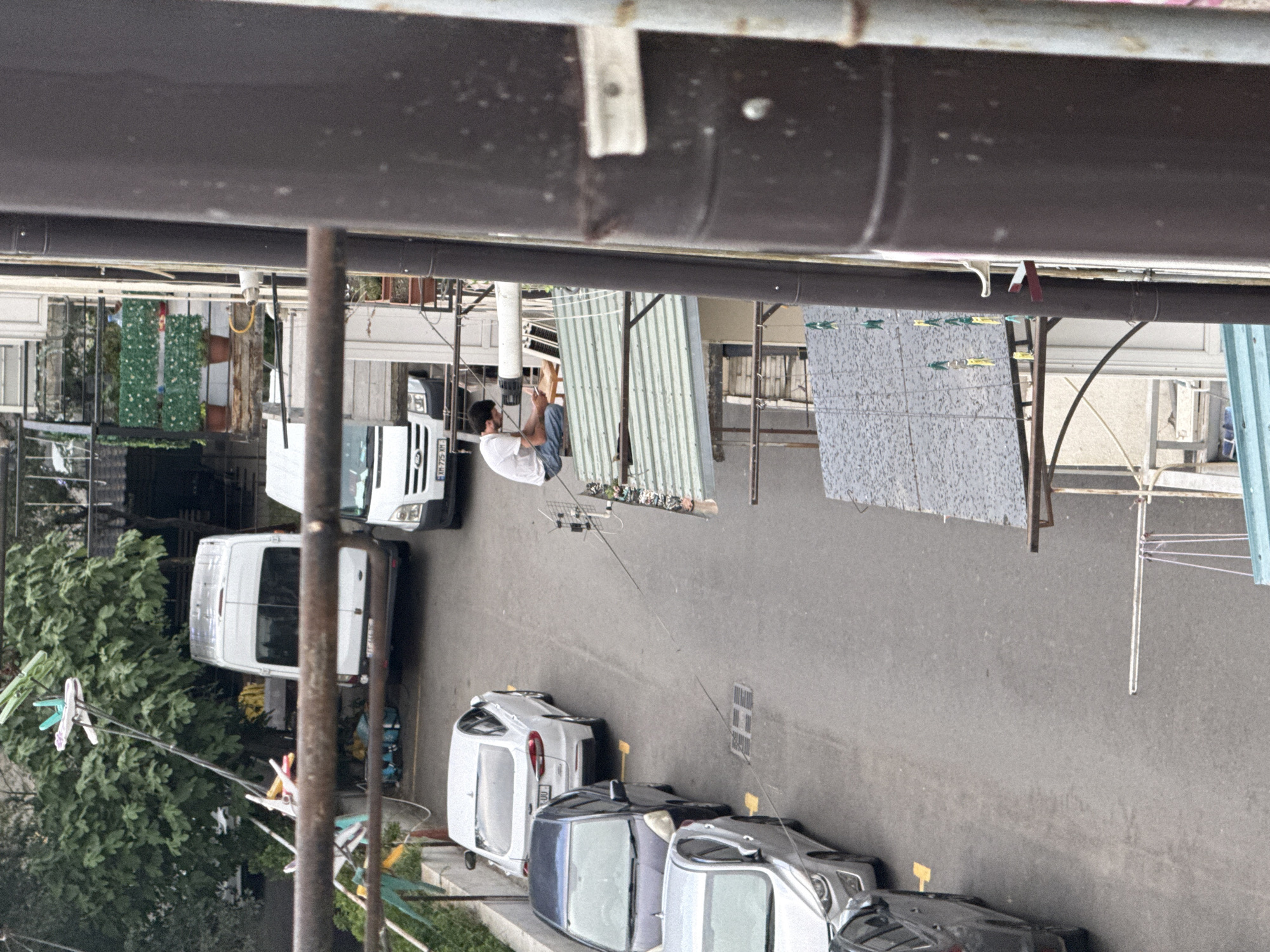In Tbilisi, Georgia, a man in a white t-shirt and blue jeans sits on a rooftop next to a white satellite dish, holding a cell phone. He is positioned on a gray, corrugated metal roof. Below the man, a clothesline with several clothespins hangs. Several cars are parked on a paved street, including a white van, a white sedan, and two dark-colored sedans. In the background, lush green trees and other buildings are visible. The scene appears to be an urban residential area.