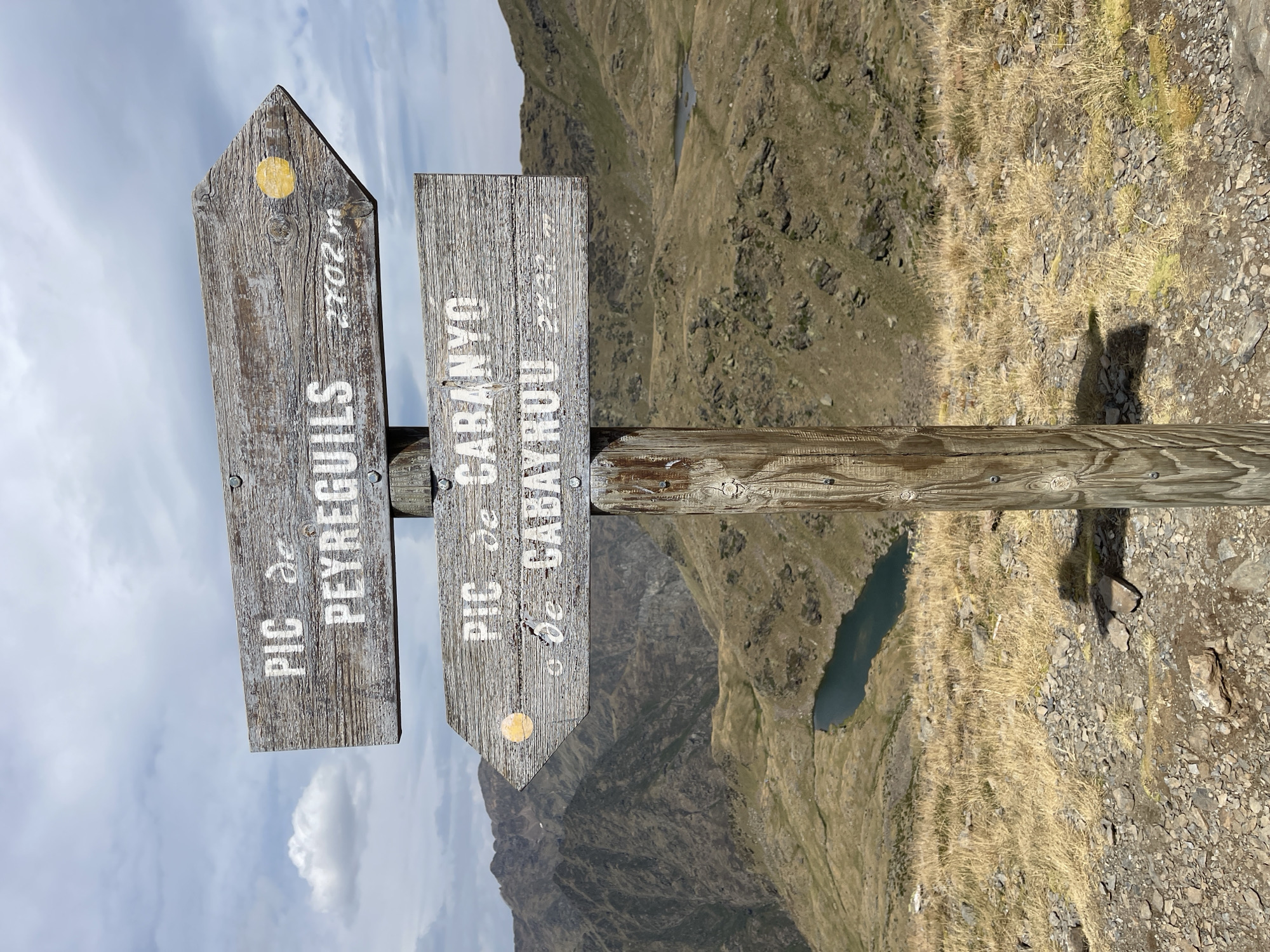 A wooden signpost with two directional arrows stands in a mountainous, arid environment. The top arrow, pointing to the right, is labeled "PIC de PEYREGUILS 2702 m". The bottom arrow, pointing to the left, is labeled "PIC de CABANYO de CABAYROU 2722 m". Both arrows have a small yellow dot near their tips. The signpost is made of weathered wood and appears to be attached to a thicker wooden post.
The background consists of rugged, brown and green mountains with a cloudy sky. A small, dark blue lake is visible in the middle ground, nestled in a valley between the mountains. The terrain around the signpost is rocky and covered with dry, golden grass. The lighting suggests it is daytime. There are no people visible in the image.