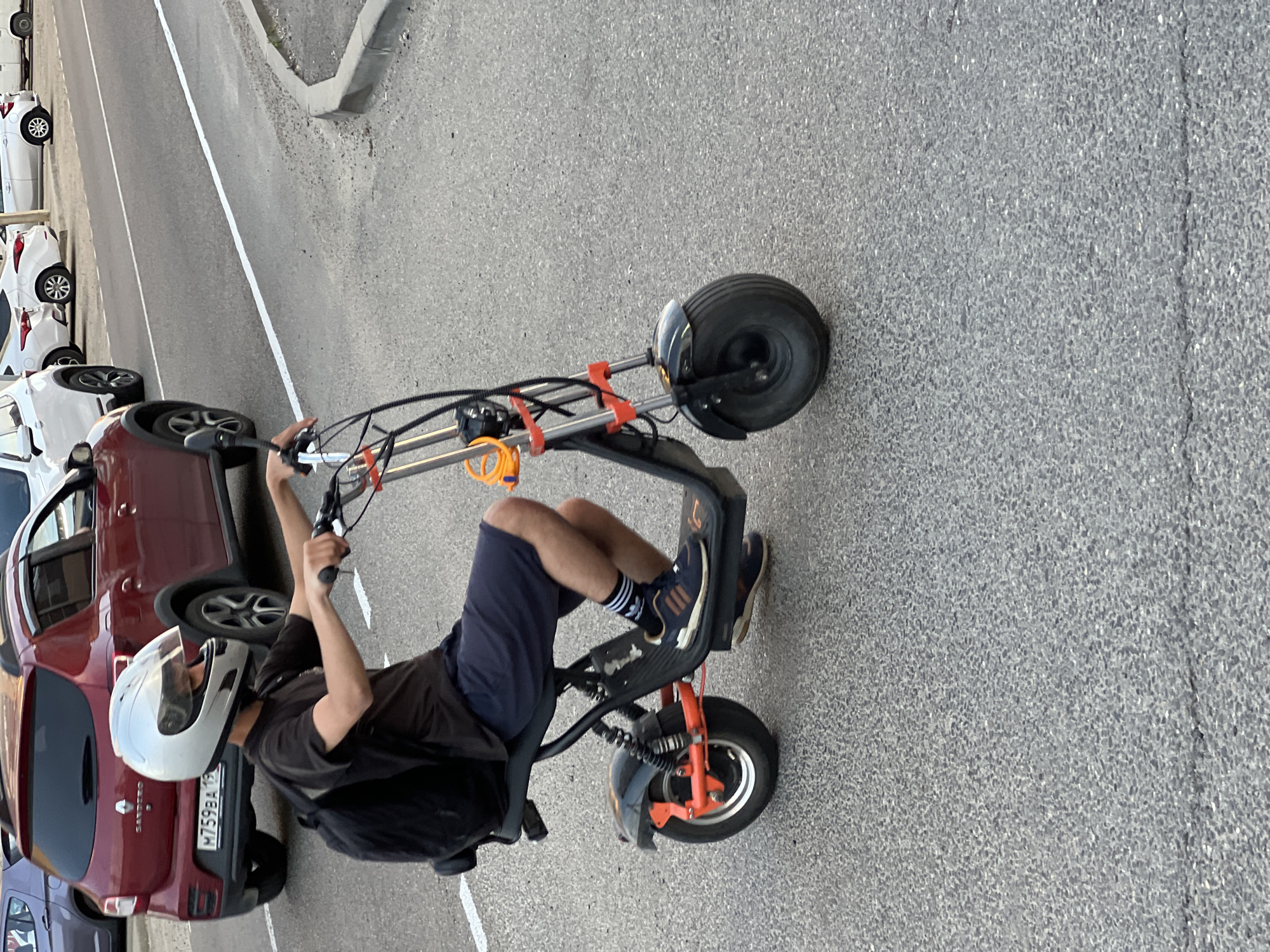 In Yablonovskiy, Russia, a person wearing a black t-shirt, dark shorts, white socks with black stripes, and navy blue Adidas sneakers rides a black electric scooter with orange accents. The rider is also wearing a silver full-face helmet. A black backpack is visible on their back. The scooter has thick tires and a suspension system. The background features a red Renault Sandero car with a license plate that reads "M759BA 10 RUS". Several other vehicles, including white cars and a white van, are parked or driving on the asphalt road, which has white lane markings and a concrete curb. The scene appears to be set on a street in a residential or commercial area.