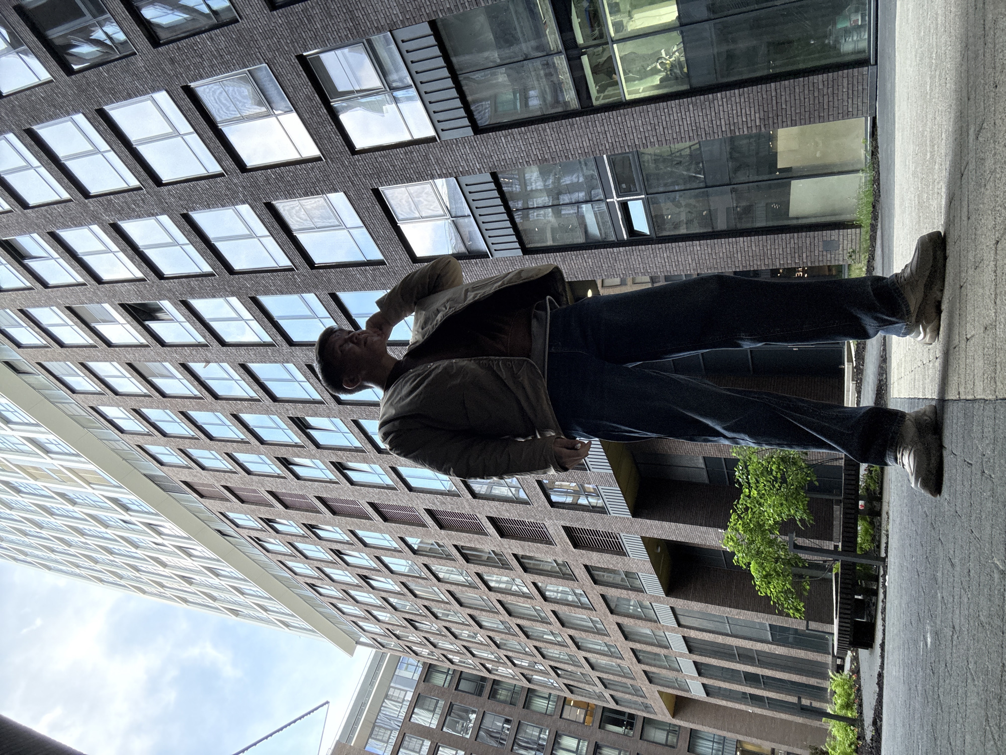 A man wearing a light-colored jacket and dark jeans stands on a gray brick walkway. He is looking up and appears to be holding his hand to his face. Behind him rises a modern brick building with numerous square windows. The sky is visible in the upper left corner of the frame. There are other structures, greenery, and reflections in the windows, adding depth to the urban scene. It is not possible to know the precise location is Moscow, Russia.
