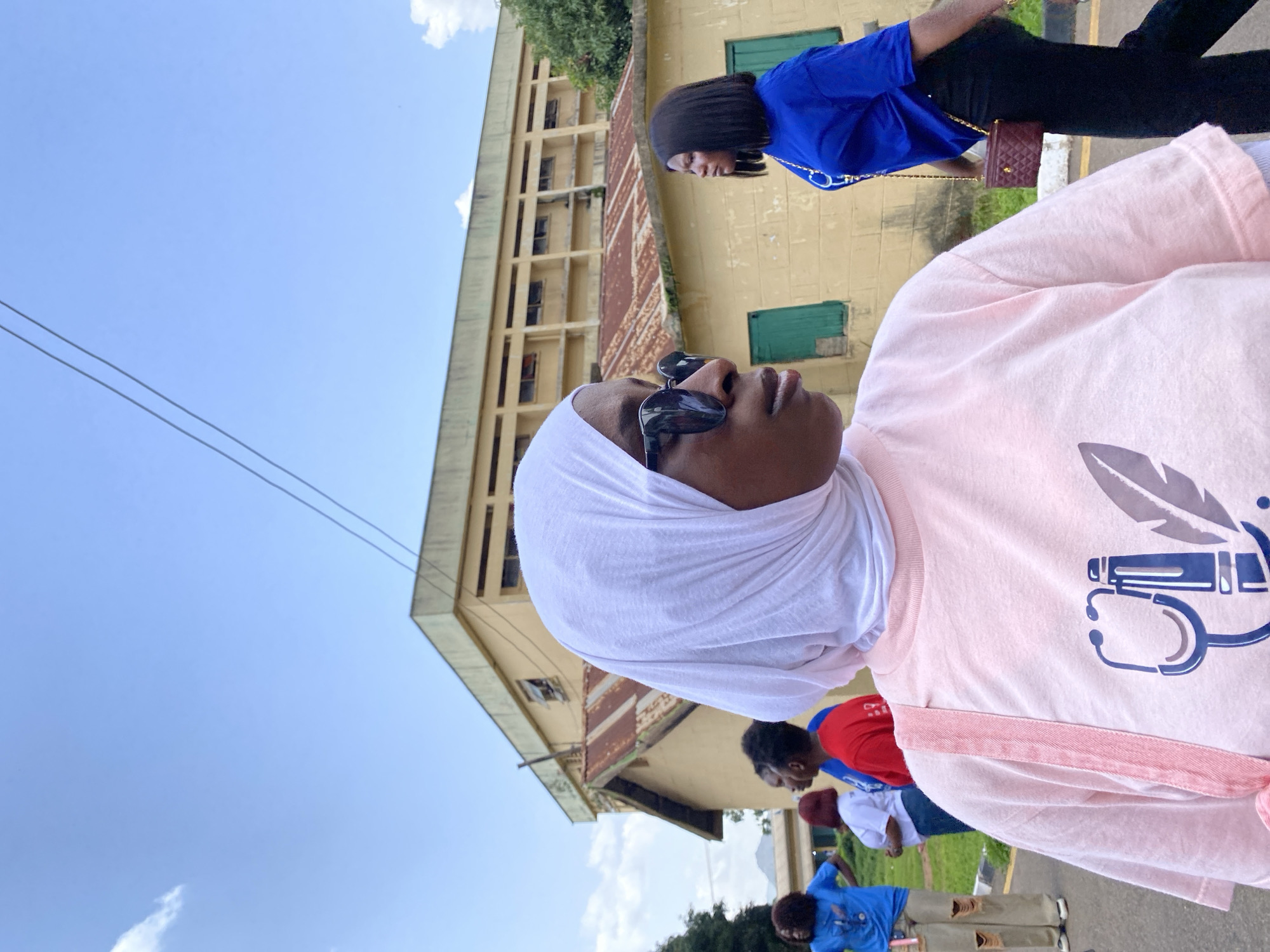 In Jos, Nigeria, on a bright day with a clear blue sky and scattered clouds, a group of women are gathered outdoors. The primary focus is on a woman in the foreground wearing a white hijab and a light pink t-shirt. She is also wearing sunglasses and looking to her right. Her t-shirt has a graphic design featuring a stethoscope and a pen. A pink strap from a bag is visible over her shoulder.
Behind her, to the left, another woman in a blue t-shirt and ripped tan pants is also looking away. Further back, a woman in a red t-shirt is visible, and next to her, a woman in a white t-shirt. To the right of the foreground subject, another woman is walking away, wearing a vibrant blue t-shirt and black pants. She is carrying a small, quilted maroon purse with a gold chain.
The background consists of a two-story building with a yellowish facade and a rusty, corrugated iron roof. The building has several windows, some of which appear broken or boarded up. Green shutters are visible on some windows. Power lines run across the top of the image. Trees and a distant hillside are visible to the left, suggesting a semi-urban or campus environment. The ground appears to be paved with asphalt.