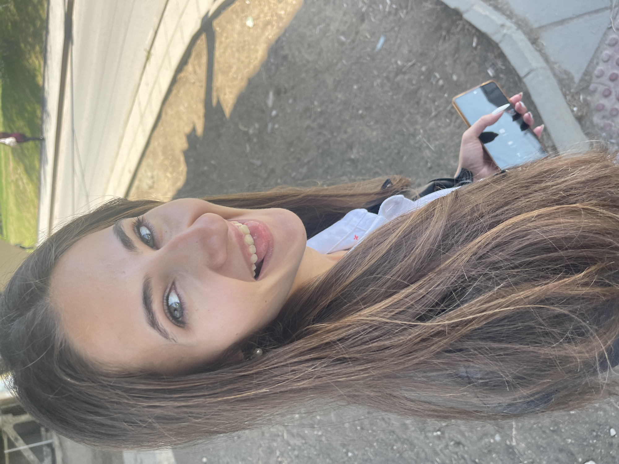 A young woman with long, straight, brown hair and captivating blue-green eyes is captured in an upward-angled shot, likely a selfie. She is smiling broadly, showing her teeth, with her tongue slightly visible, conveying a cheerful and playful expression. She has fair skin and appears to be wearing a white top, possibly with subtle red embroidery on the chest area, beneath a dark outer garment like a jacket or coat.
In her right hand, she holds a smartphone with a gold or bronze-colored frame and a dark, reflective screen. Her fingernails are long and painted white. The setting is an outdoor urban environment in Sofia, Bulgaria. The background features a paved surface, resembling a sidewalk or pathway, adjacent to a curb and what appears to be a road. The ground exhibits variations in texture and color, with lighter concrete and darker, possibly earthy, patches. A patch of green grass is visible on the far left. Strong, distinct shadows are cast across the ground, indicating bright sunlight and suggesting the time of day is either morning or afternoon. The weather is clear and sunny. No visible text is present on any objects or surfaces in the image.