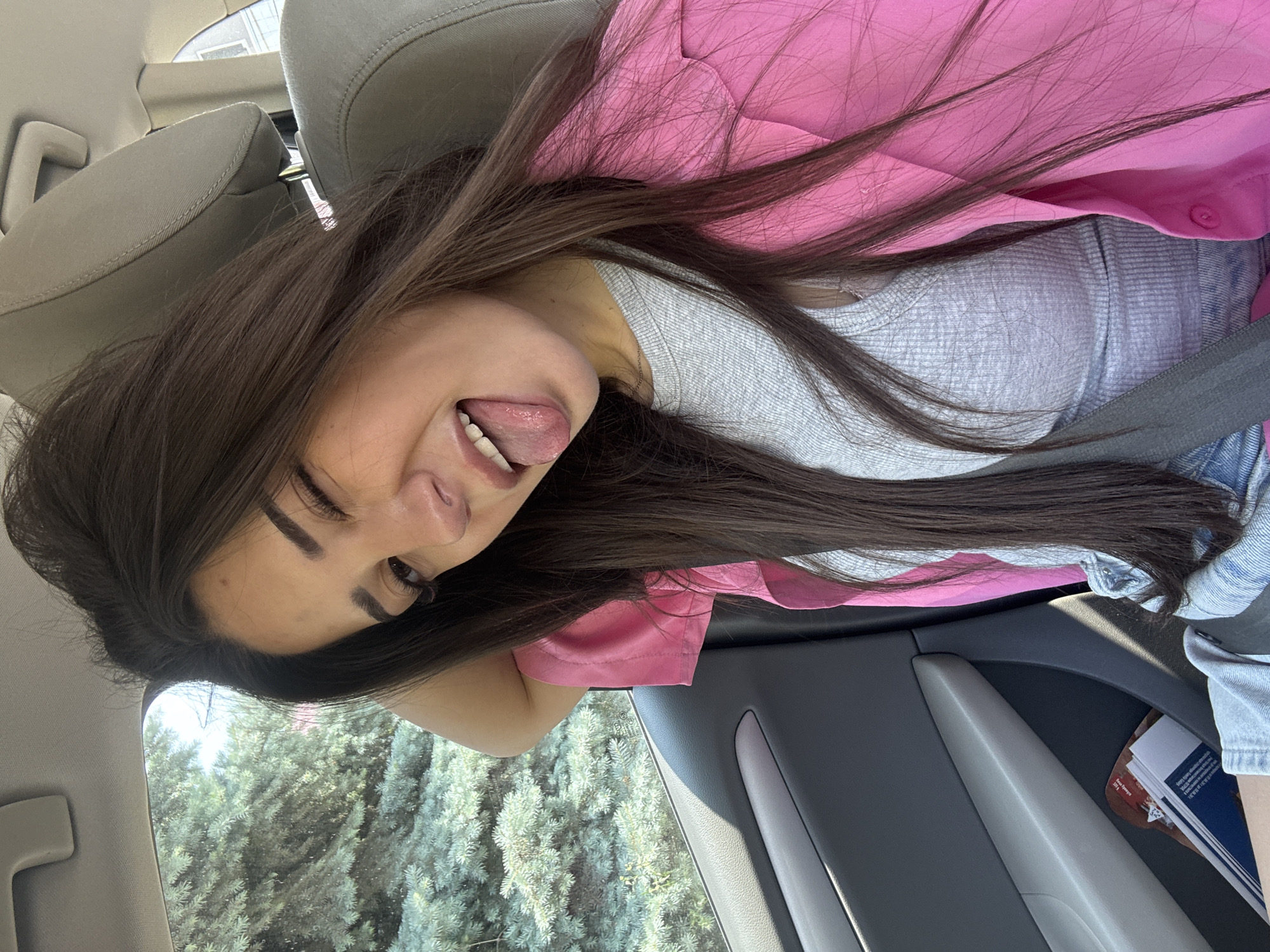 A young woman with long, dark brown hair is captured in a playful pose inside a car. She is looking towards the camera with a cheerful expression, sticking her tongue out. Her hair is spread across the car seat and her shoulders. She is wearing a light grey ribbed long-sleeve top, a bright pink jacket or shirt over it, and what appears to be denim clothing. A dark grey seatbelt is visible across her chest.
The setting is clearly the interior of a car, likely the front passenger seat, indicated by the beige-colored headrest and part of the dashboard. Through the car window, dense evergreen trees are visible, suggesting an outdoor, possibly natural or landscaped environment, under bright daylight.
Notable details include her well-defined eyebrows, visible eyelashes, and the car's interior design, including a grey door panel with a handle. In the door pocket, several papers are stacked; a blue spine with the partial text "202..." is faintly visible on one, but other text is illegible. The image is oriented sideways, with the woman's head tilted to the right, and the external scenery on the left. The overall mood conveyed is lighthearted and fun. The location is Plovdiv, Bulgaria.