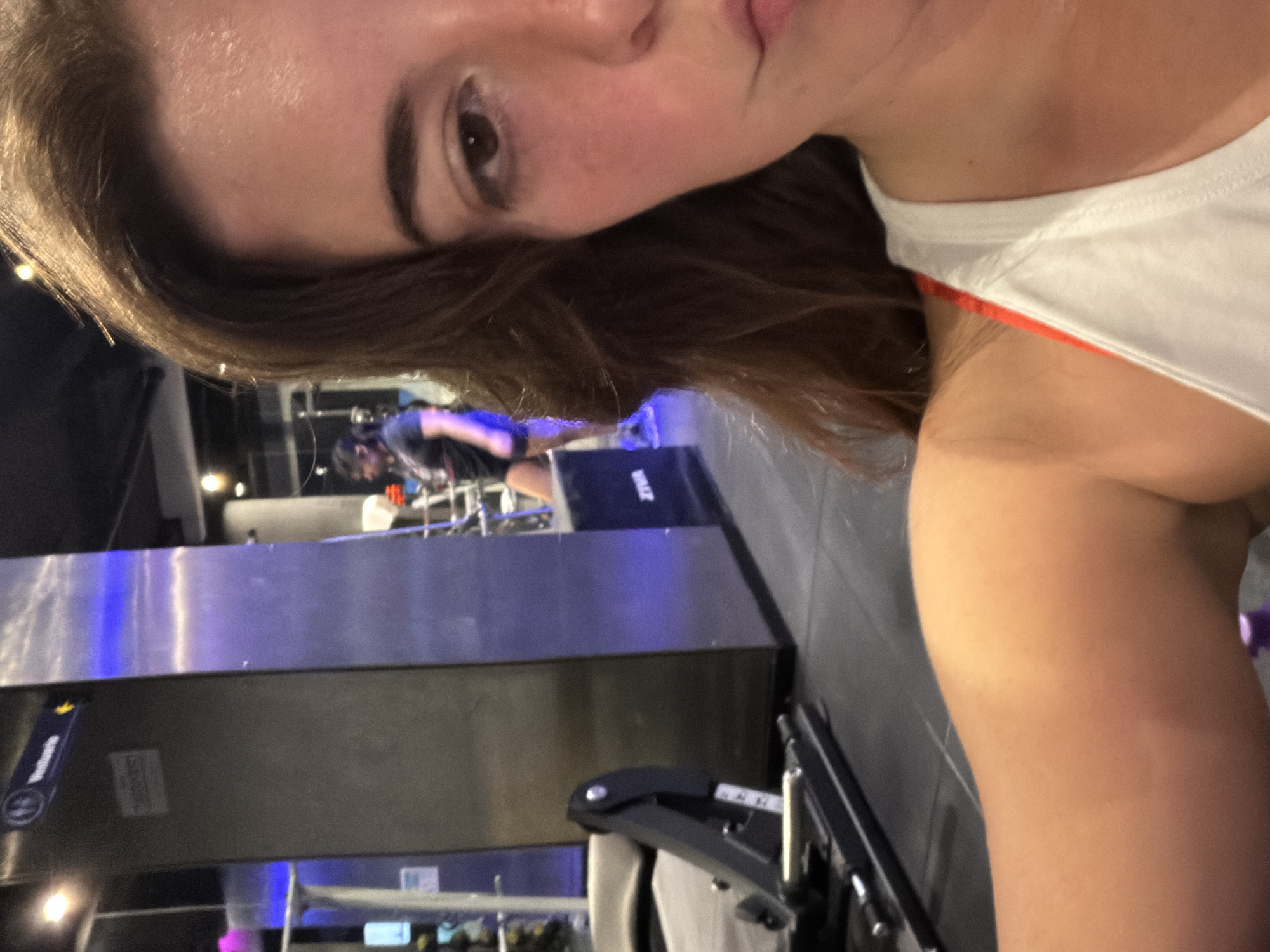 The image shows a selfie of a woman with brown hair. She is wearing a white tank top with an orange accent. The background appears to be a gym or fitness center, with exercise equipment visible. There are other people working out in the background. There is a sign with the word "Salidas", likely indicating the exits. The presence of "Salidas" may indicate this image was taken in a bus terminal in Buenos Aires, Argentina.
