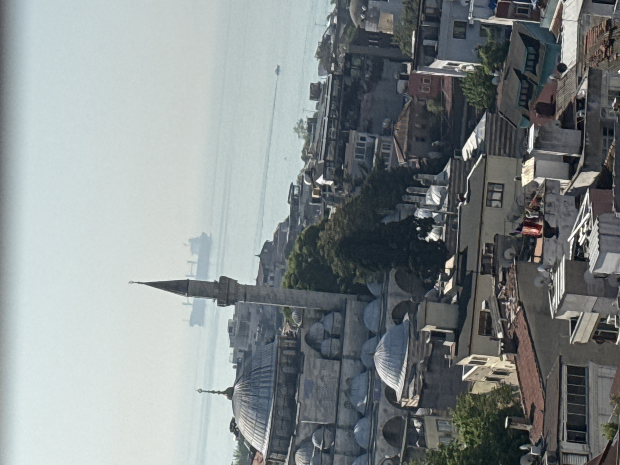 The image shows an aerial view of an urban landscape with a prominent mosque in the foreground. The mosque features a series of domes and a minaret. The environment includes a body of water, possibly a sea or large river, with ships visible on the horizon. The cityscape is densely packed with buildings of varying heights and architectural styles. The overall tone is slightly hazy, suggesting a mild weather condition.