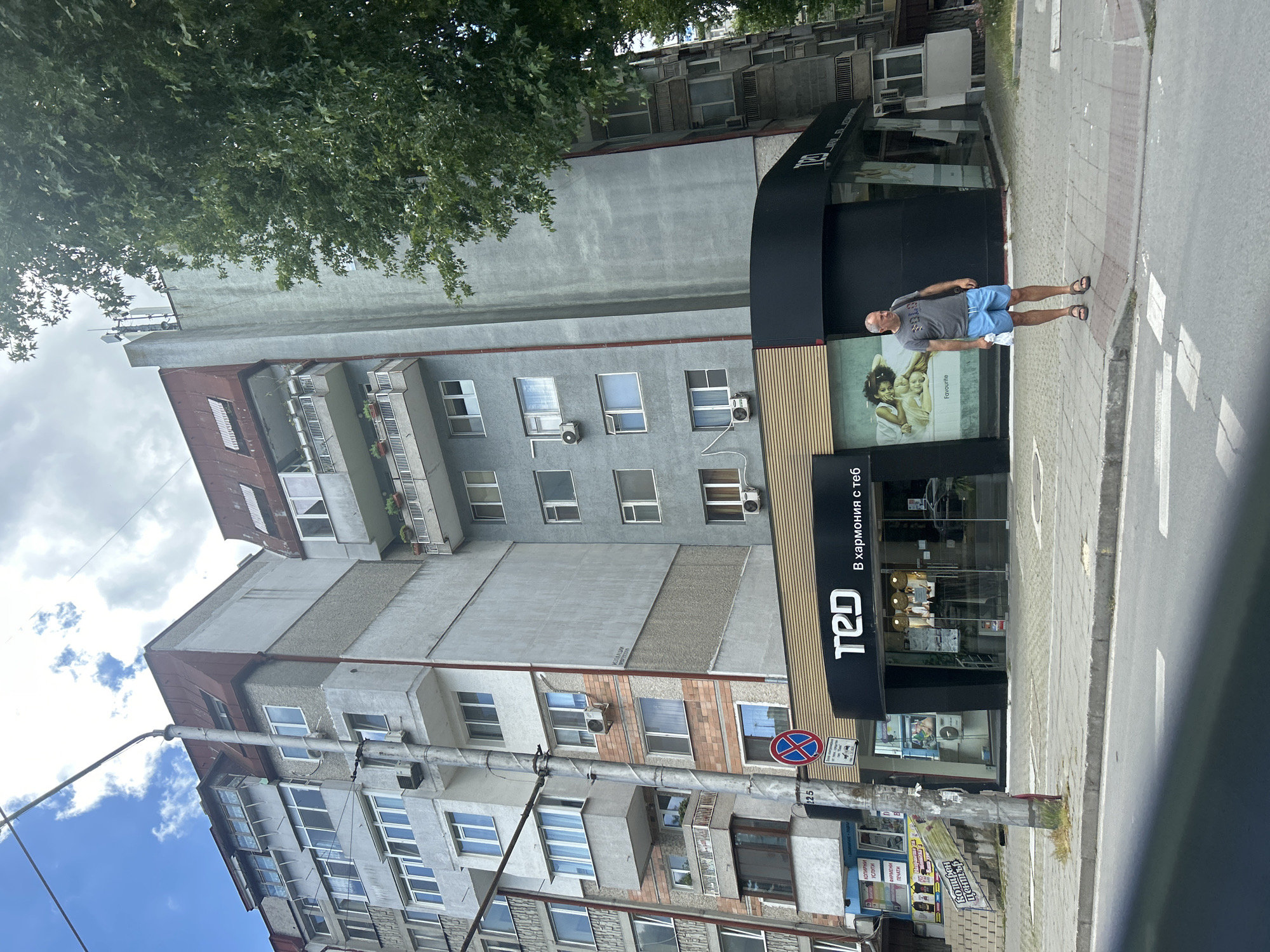 The image shows a street scene in Varna, Bulgaria, likely in front of a retail establishment with a black facade and a prominent logo reading "TED". A middle-aged man, wearing a grey t-shirt and blue shorts, stands on the sidewalk. He is holding a white bag in his right hand and appears to be looking to his right. Behind him, the building features multiple stories with windows and balconies, typical of residential architecture in the area. A traffic sign with a red circle and a blue cross is visible on a pole next to the building. The street in the foreground is paved, and there are some trees and greenery in the background. The sky is partly cloudy.