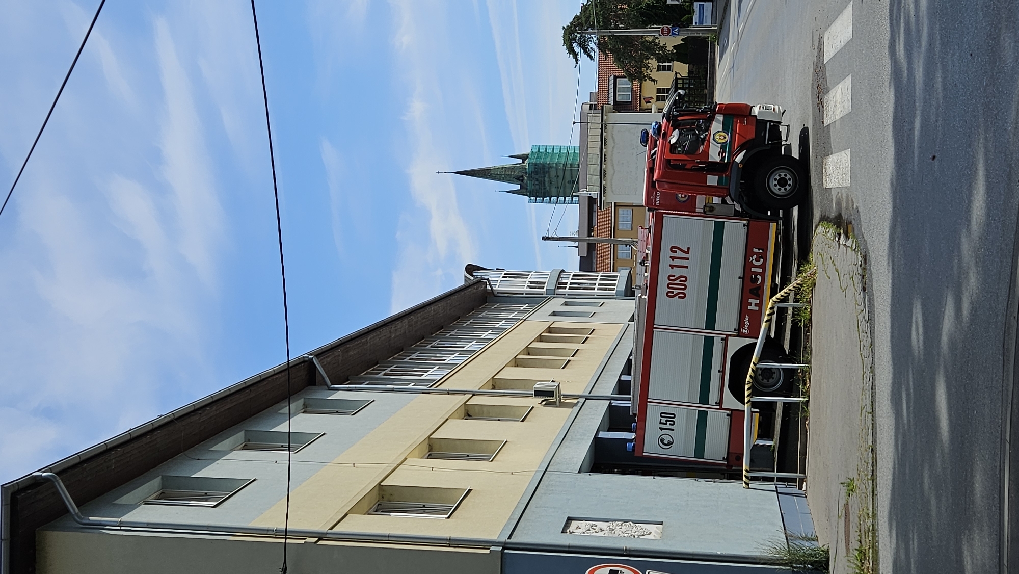 An outdoor, eye-level, medium shot captures a red and white fire truck parked beside a building in Prešov, Slovakia. The building, painted in light blue and yellow, features a row of windows along its facade. The fire truck, identified with "150" and "SOS 112" markings, is positioned in front of a garage entrance. In the background, a church steeple, partially covered in green scaffolding, rises against a blue sky dotted with white clouds. The street in the foreground includes a crosswalk.