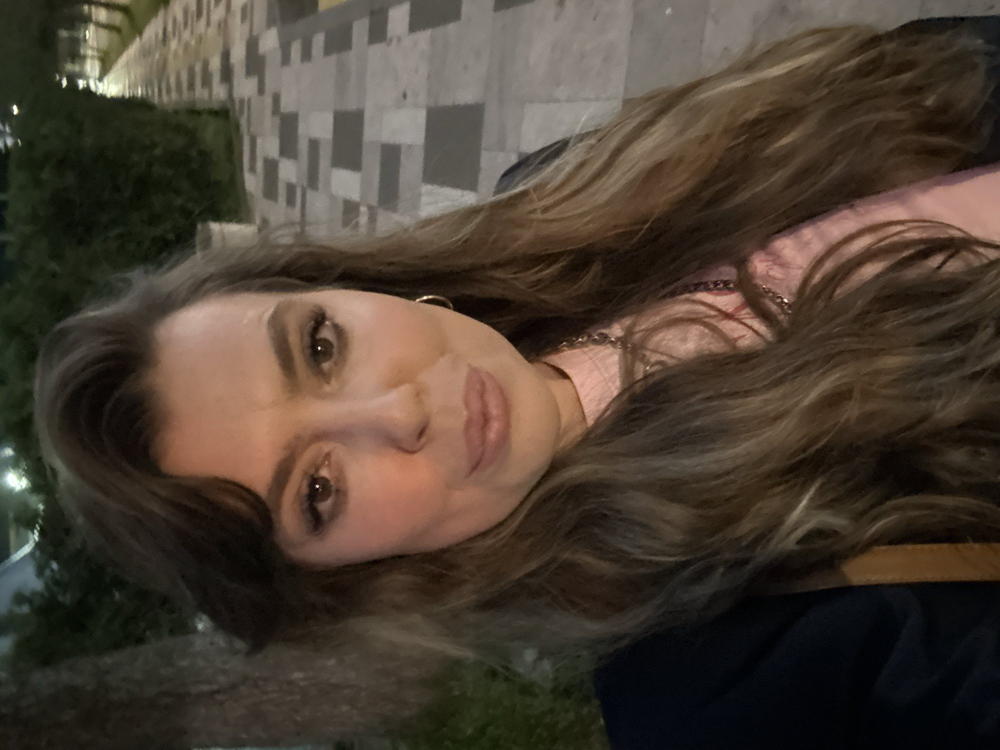 The image is a selfie of a woman with long, wavy brown and blonde hair, taken at night in Batumi, Georgia. She is wearing a light pink shirt, a dark-colored jacket, and gold earrings. A gold chain necklace is visible on her neck. The background shows a tiled walkway and greenery, consistent with an urban environment. The lighting suggests it's evening or night, with artificial lights illuminating the scene. The architecture in the background is modern and includes buildings with geometric designs. The photo appears to be taken from a slightly low angle.