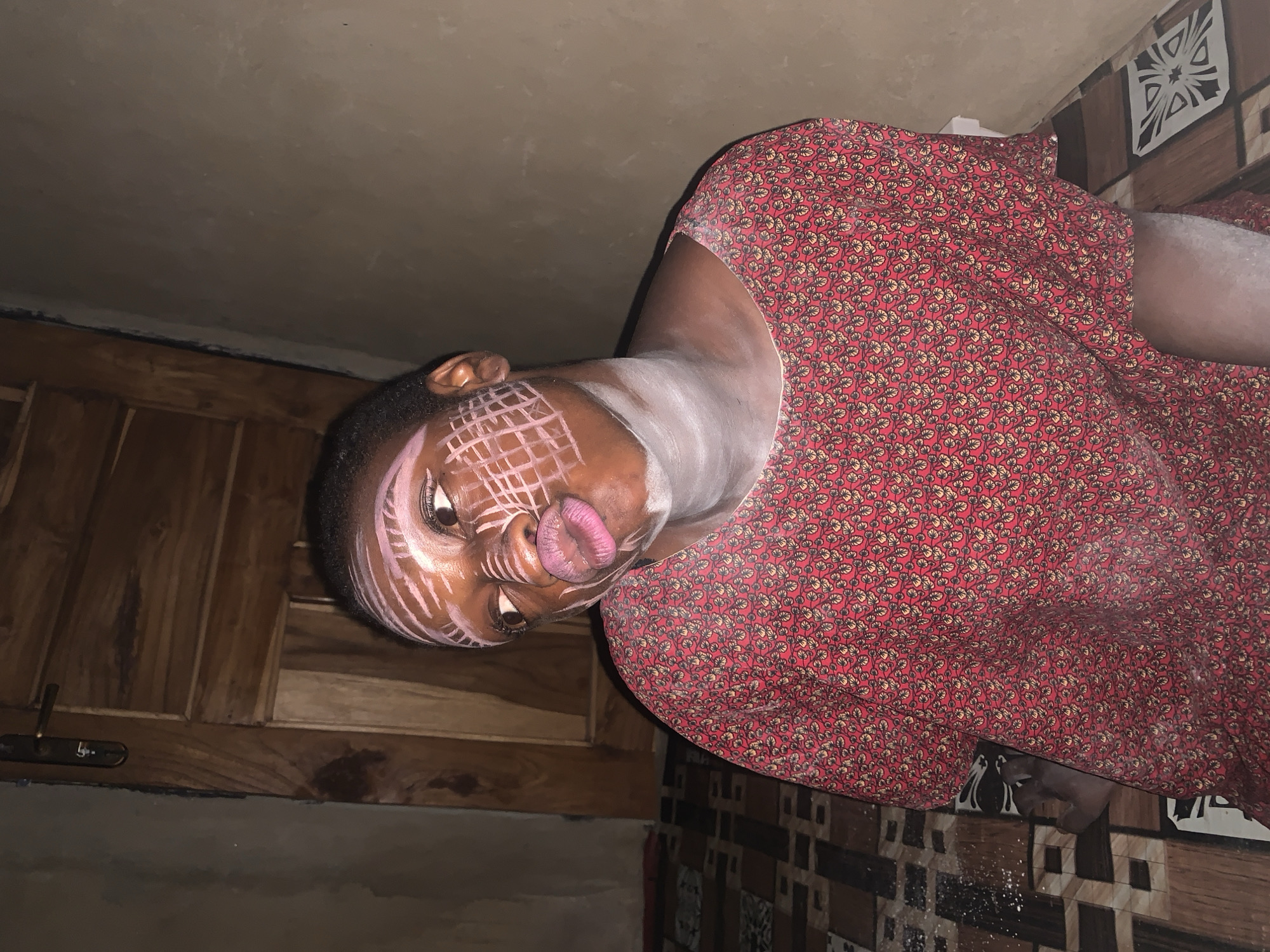 A young person with dark skin and a red, patterned dress is seated indoors. The person has white paint or makeup applied in a grid pattern on their face and a white band around their neck. Their lips are pursed and painted pink. The background includes a wooden door with a metal handle, a wall, and a patterned tiled floor. The scene appears to be within a residential setting in Agban, Nigeria.