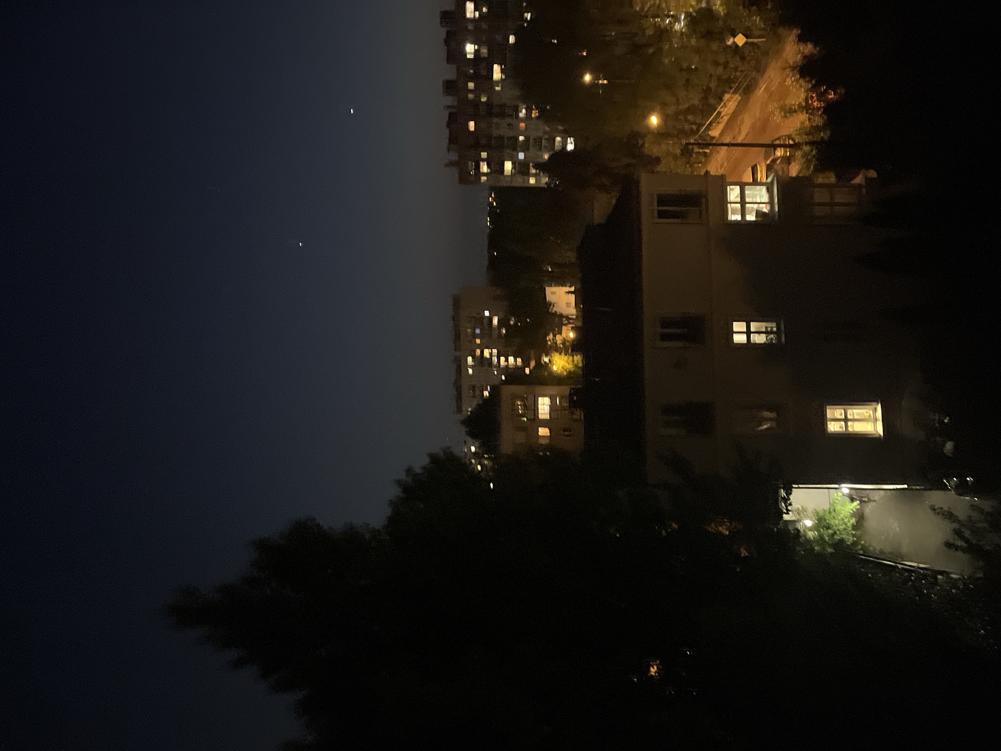 The image depicts a night scene in an urban area. Several buildings with lit windows are visible in the middle ground, with denser apartment blocks further in the background. Streetlights illuminate the area. A dark, silhouetted tree dominates the foreground. Two small bright dots are visible in the upper left of the image, appearing to be stars in the night sky.
