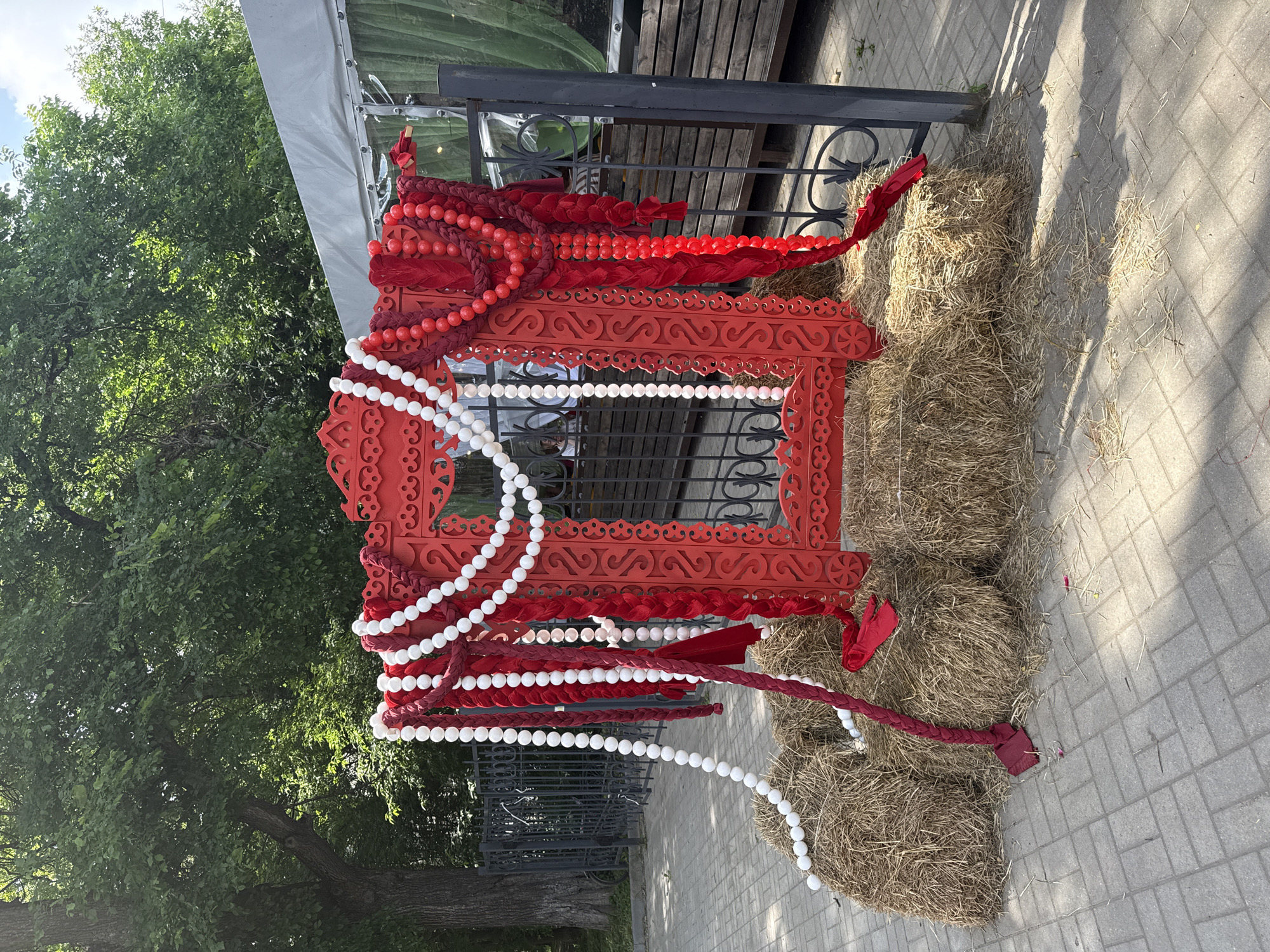 A vibrant red, ornately carved wooden frame, decorated with strings of white and red beads and thick red braided fabric, is set up outdoors on a paved area in Vologda, Russia. The frame rests on several large bales of straw. Behind the frame, a dark metal fence and lush green trees are visible. To the right of the frame, part of a white tent structure and wooden decking can be seen, suggesting a public event or outdoor seating area. The lighting indicates it is daytime, with shadows cast on the pavement. No people are clearly visible within the frame or immediately surrounding it.