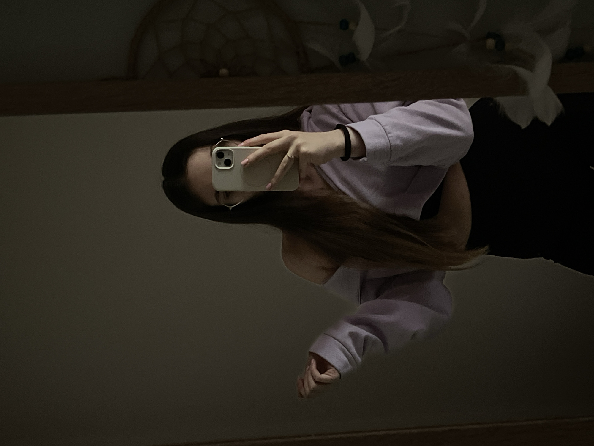 A woman is taking a mirror selfie with her phone. She is wearing glasses, a lavender sweatshirt, and a black bracelet. Her hair is brown. The background is a dark wall. Above the mirror is a decorative object. The location is unknown.